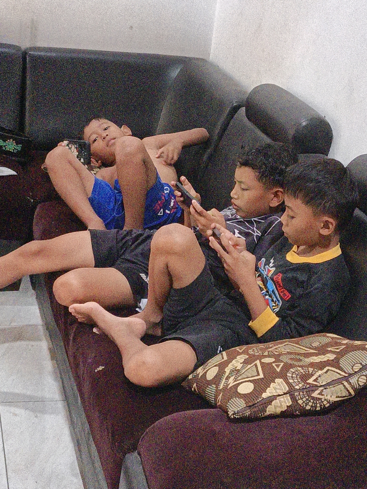The image shows three young boys lying on a dark brown sofa, engaged with their mobile phones. The boy on the far left is lying on his back, propped up on his elbows, wearing blue shorts and holding a phone in his left hand. In the center, a boy is also lying on his back, looking down at a phone in his hands and wearing a black shirt with a white design. To the right, another boy is lying on his side, also focused on his phone and wearing a black shirt with yellow accents. The sofa is a deep brown, with some visible cushions. The background appears to be a plain, light-colored wall. The overall impression is a casual, indoor scene in what is likely a home environment.