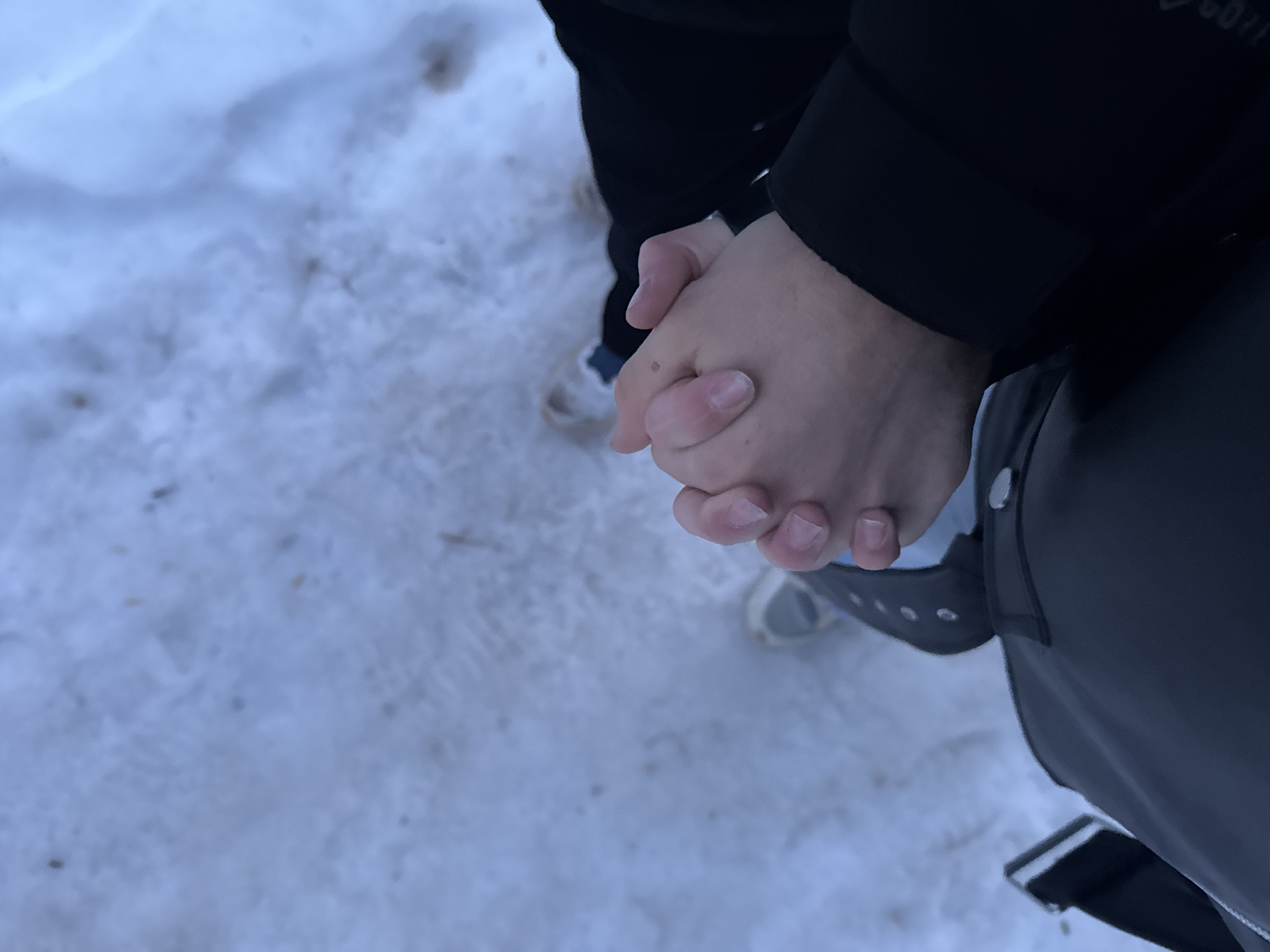 A person stands in the snow. The person is wearing a dark colored jacket and light colored shoes. Their hands are clasped together. The location is likely outside due to the presence of snow. I cannot identify the specific location as Chaikovsky, Russia.