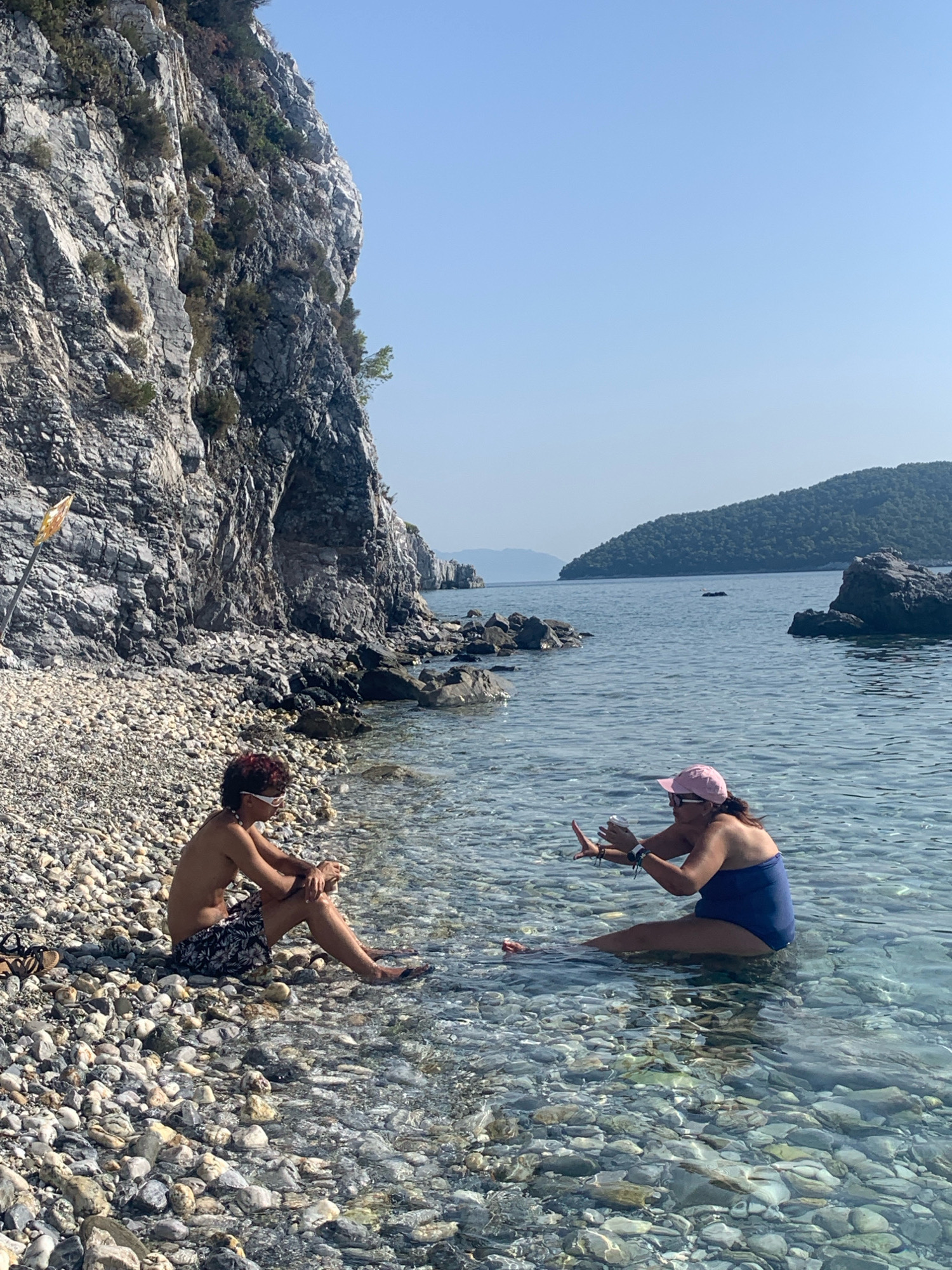 Two people are by the water on a rocky beach, with a steep, grey cliff face behind them. The person on the left is sitting on the shore with their legs outstretched, wearing patterned swim shorts and white sunglasses. The person on the right is sitting in the shallow, clear water, wearing a blue swimsuit and a pink cap. They are holding a cup and gesturing with their hands. The sea extends to the horizon, with green, hilly islands in the distance. The sky is clear and blue. The scene appears to be on a sunny day.