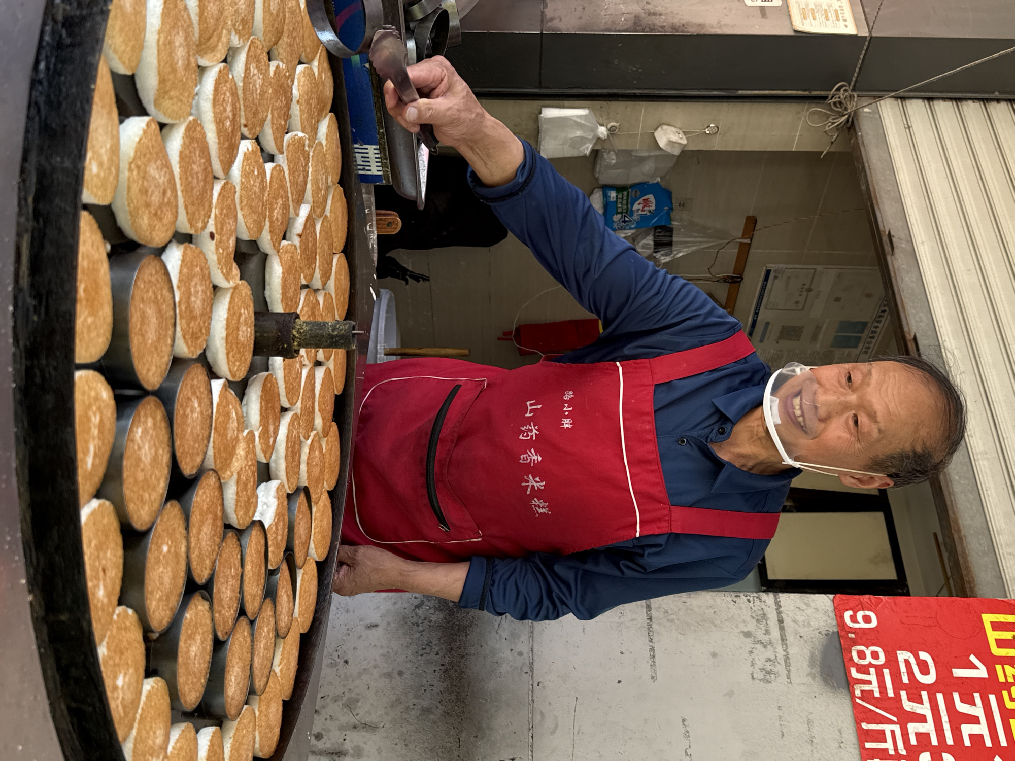 An eye-level, indoor shot captures an elderly Asian man in what appears to be Beibei, China, standing behind a vertical cooking apparatus, smiling at the camera. The man wears a long-sleeved blue shirt, a red apron with white trim, and a clear plastic face shield. The apron has Chinese characters on it.
The cooking device is a large, black, rotating circular metal container filled with cylindrical food items. These items are white on the outside and tan on the inside, and they are arranged in a grid. The man holds a long metal spoon or spatula in his right hand, which he uses to turn the items.
To the right of the man, a red sign with Chinese characters and numbers (1, 2, 9.8) stands on the ground. Behind him, a partially open metal shutter covers a doorway. Within the doorway, a dimly lit space holds a box of tissue and other miscellaneous objects. Above the doorway, a sign and electrical wiring can be seen. The floor area is light grey and appears to be concrete.