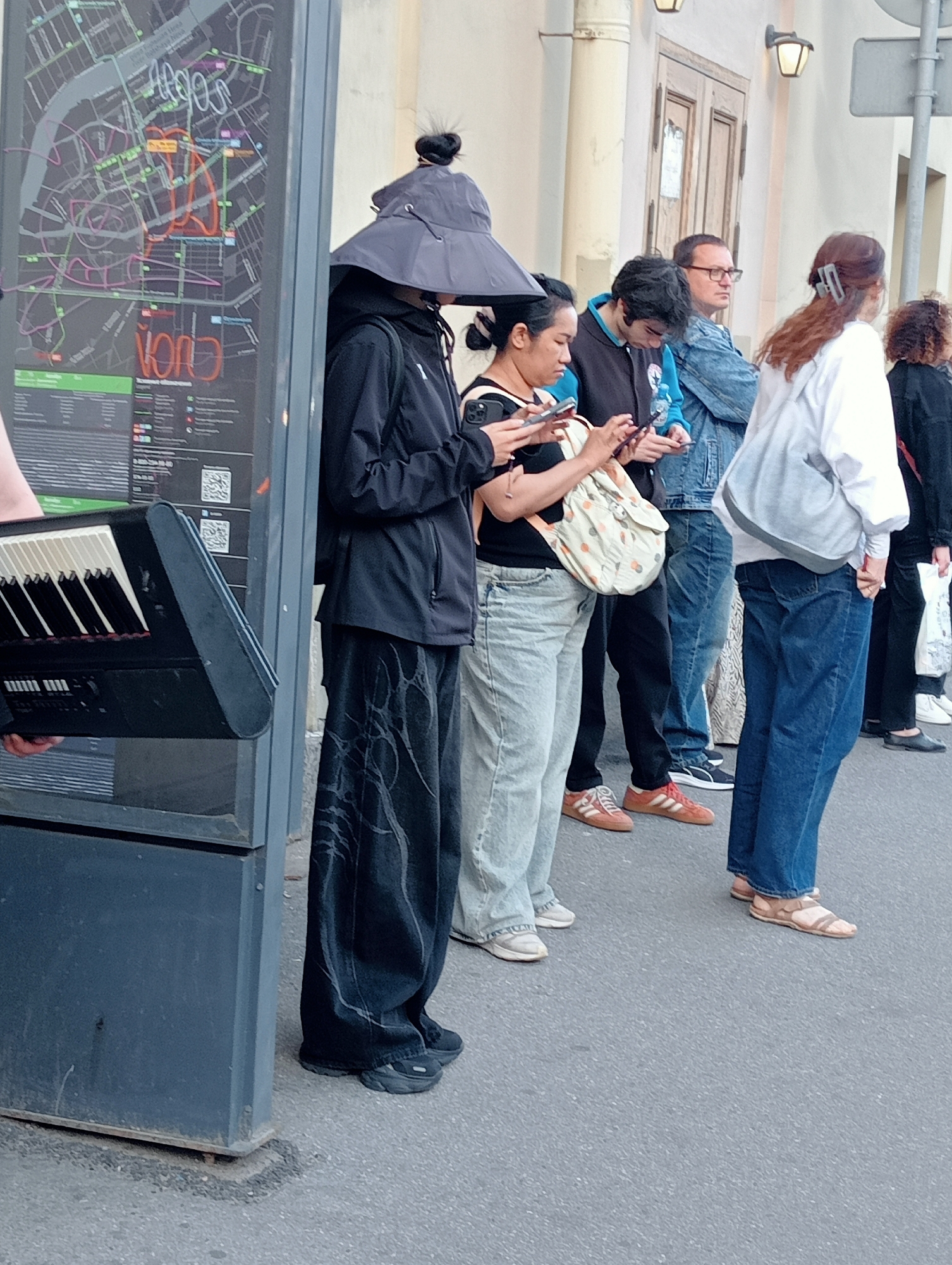 In a street scene set in Saint Petersburg, Russia, a group of people stand in line, their attention directed towards their mobile phones. The setting appears to be outdoors, with a building featuring classical architectural elements in the background. A large informational map is mounted on a dark grey kiosk to the left of the frame, displaying a city map with various routes and landmarks marked in different colors. The people are dressed casually; one individual in the foreground wears a wide-brimmed hat and a black outfit, obscuring most of their face. Another person holds a portable electronic keyboard. The overall mood seems neutral, with individuals engrossed in their devices. The lighting suggests it is daytime.