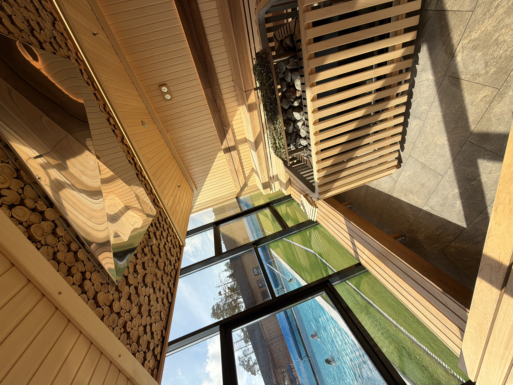 The image shows the interior of a sauna located in Votkinsk, Russia, adjacent to an outdoor swimming pool. The sauna is constructed primarily of wood. The interior walls and ceiling are lined with light-colored wooden planks. One wall features a decorative panel made of circular wood cuts. A stainless steel, pyramid-shaped fixture is mounted on this wall. The sauna includes a window view of the outdoor pool, artificial turf surrounding the pool, and a building across the pool. A wooden sauna heater filled with stones sits in one corner. The floor outside the heater is tiled. Sunlight enters the sauna. Reflected in the glass of the window are trees and a building. Swimmers are visible in the pool.