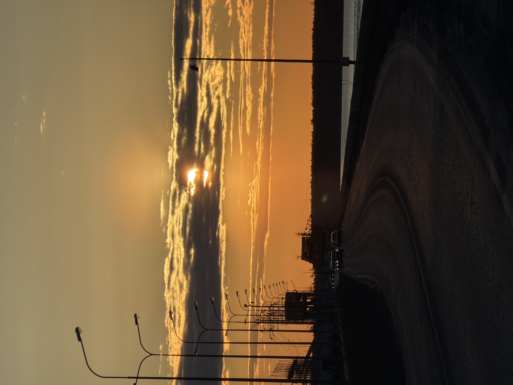 The image shows a sunset view in Chaikovsky, Russia, next to the Kama River. The setting sun is partially obscured by clouds. A highway runs along the right side of the image with at least three vehicles travelling on it. A line of streetlights runs along the side of the road. The sky is yellow and orange near the horizon, transitioning to a lighter yellow as it rises. In the background, a power plant or industrial facility is visible. High voltage towers and lines crisscross the area beyond the streetlights.