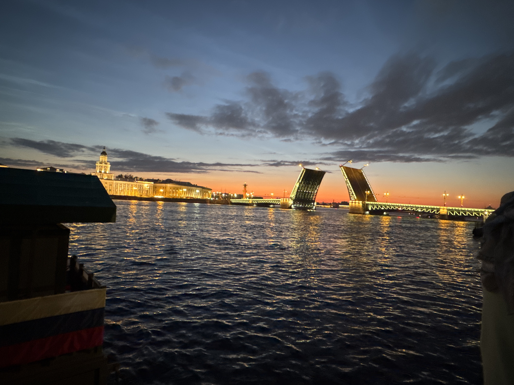 The image depicts a city skyline at twilight, likely St. Petersburg, Russia, given the prominent drawbridge. The sky is a gradient of deep blue to orange, with scattered clouds. The water in the foreground is dark and rippled, reflecting the lights from the city. On the left side of the frame, a dark structure, possibly a boat or pier, with a flag of Russia is visible. In the background, a long building with many illuminated windows stretches across the horizon, topped by a dome. Two sections of a drawbridge are raised, creating a dramatic silhouette against the colorful sky. The bridge is lined with lights, and its structure is clearly visible. The overall scene evokes a sense of calm and grandeur, characteristic of a waterfront city at dusk.