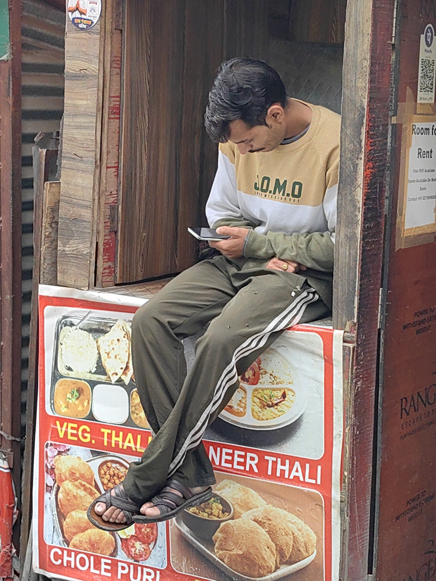 A young man with dark hair and a mustache is seated in an open doorway of a wooden structure, looking down at his smartphone. He is wearing a beige and white long-sleeved shirt with the letters "J.O.M.O." on it, and dark green track pants with white stripes. His feet, clad in dark brown sandals, rest on a red and white sign advertising food items, including "VEG. THALI" and "CHOLE PURI". The sign also displays images of various Indian dishes. The environment appears to be an outdoor setting in Vashisht, India, possibly a food stall or small shop, with wooden planks and metal framing visible. A faded QR code and some text are affixed to the right side of the doorway, possibly for payments or advertisements. The lighting suggests it is daytime.