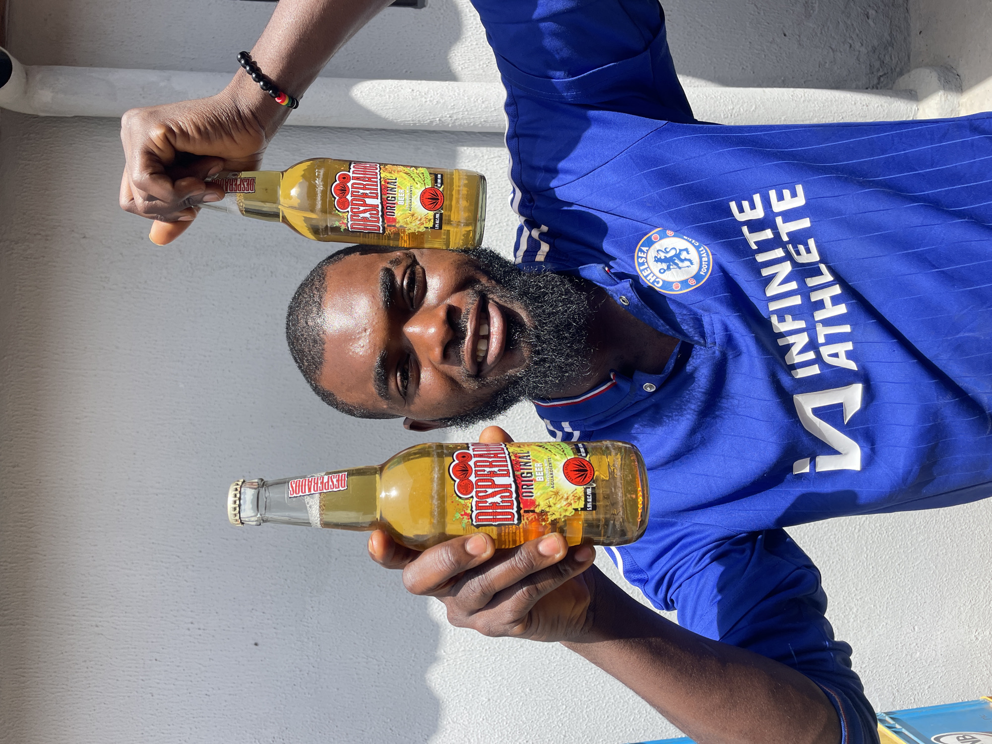 The image shows a man holding two bottles of Desperados beer. He is wearing a blue Chelsea Football Club jersey with the words "INFINITE ATHLETE" printed in white on the front. The man has dark skin, a beard, and is smiling at the camera. He is also wearing a bracelet with black, red, and green beads. The bottles of beer are clear glass with yellow liquid and feature the Desperados brand name and logo, which includes apples and chili peppers. One bottle is being held in the man's right hand, and the other is positioned above his head in his left hand. The background is a textured white wall, suggesting an outdoor setting, possibly in sunlight given the strong shadows. The overall impression is one of a casual, celebratory moment. The visible text confirms the brand of beer and the sports team the man is supporting. The scene appears to be taking place during the daytime, and the bright lighting suggests fair weather. The image likely captures a moment of leisure or enjoyment.
