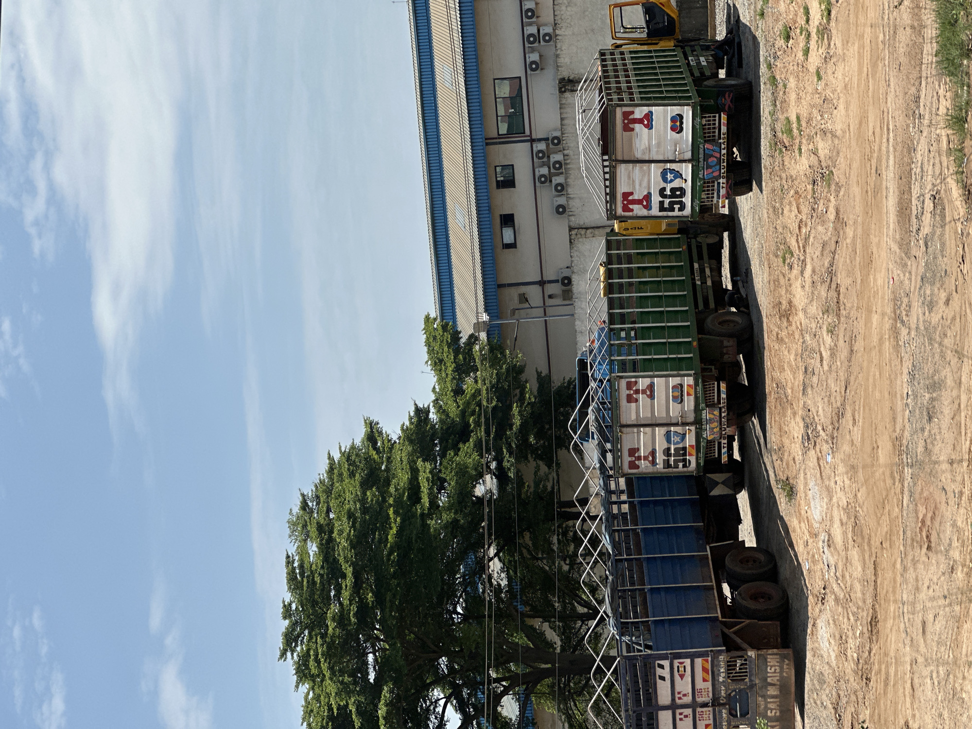 The image depicts a lot with several trucks parked on a dusty, uneven surface. In the background, a light-colored building with a blue roof and multiple outdoor air conditioning units is visible. To the left of the building, a large, leafy green tree stands prominently.
Three trucks are the primary subjects in the foreground. The leftmost truck has a blue cargo container with a metal grate enclosure. The middle truck has a green cargo container with a similar metal grate enclosure, and the rightmost truck, also with a green cargo container and metal grate, has its driver's side door open. This truck has "T 569" prominently displayed on its back, along with other colorful markings. The truck to its left has "56" repeated on its cargo area. The truck on the far left has text on its rear bumper, partially obscured, that reads "KI SAI MAISHI CITY". Another truck in the middle has "AL-BARIKA" and "TI OLUWA LASE" visible on its rear.
The ground is predominantly dirt and sand, with patches of sparse green grass. The sky above is a bright blue with scattered white clouds. The overall scene suggests an industrial or logistical area, possibly a transportation hub or a warehouse loading zone. The location is identified as Magboro, Nigeria.