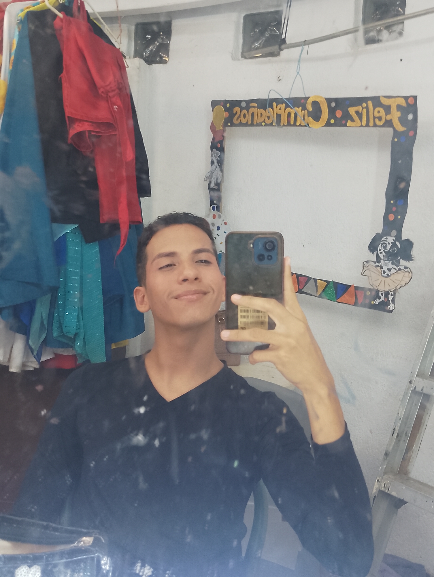 An image captured in Porlamar, Venezuela, depicts a young adult male taking a mirror selfie. The subject, positioned centrally, has short dark hair and wears a black long-sleeved V-neck shirt. He holds a dark-colored smartphone, equipped with a multi-lens camera module, in his right hand, angled to capture his reflection and the surrounding environment. A barcode sticker is visible on the rear of the phone or its case.
The background to the left of the subject displays various articles of clothing or costumes, predominantly in red, blue, black, and teal with sequined embellishments, hanging from visible hangers. The wall is white. Above, two rectangular dark openings or vents are mounted high on the wall, with a horizontal metal bar positioned below them, from which a blue string hangs.
To the right, a dark-colored, decorated frame, likely a prop for photos, is visible. It features gold lettering that reads "Feliz Cumpleaños" (Happy Birthday) in a mirrored orientation. The frame is adorned with colorful dots and a cutout figure of a black and white spotted dog, possibly a dalmatian, wearing a light brown tutu. A string of multi-colored triangular flags is also attached to the lower right section of the frame. On the far right, a silver-colored metal stepladder leans against the white wall. The lower portion of the reflective surface appears smudged or dusty, with multiple small white spots.