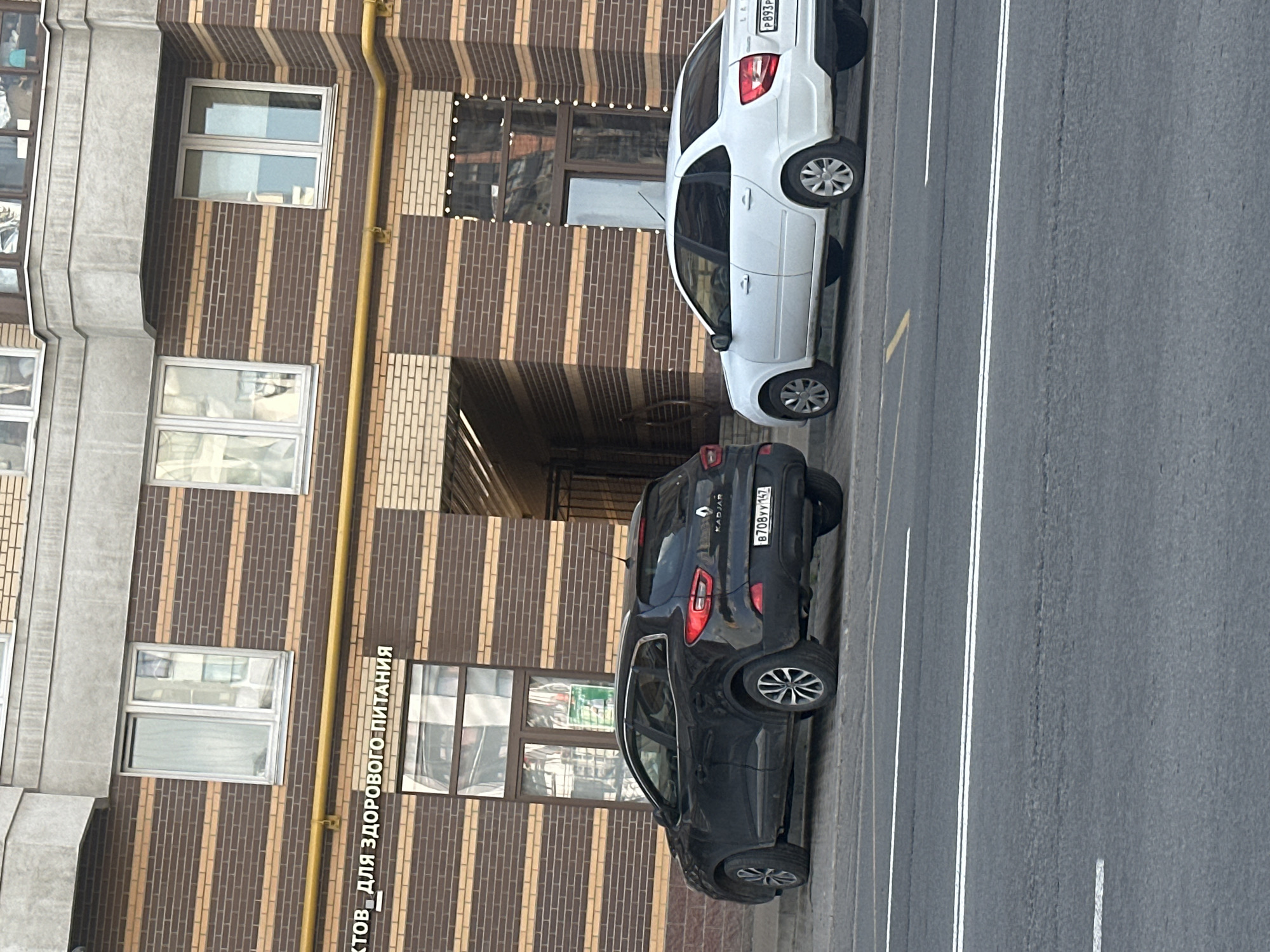 A black Renault Kadjar and a white sedan are parked on the side of a street in front of a brick building with horizontal beige stripes. The black car is on the left and the white car is on the right. The black car has a license plate that reads "B708VV 147". The white car has a license plate that reads "P893P". The building has several windows and an arched entryway. The street has white lane markings. The sky is not visible in the image.