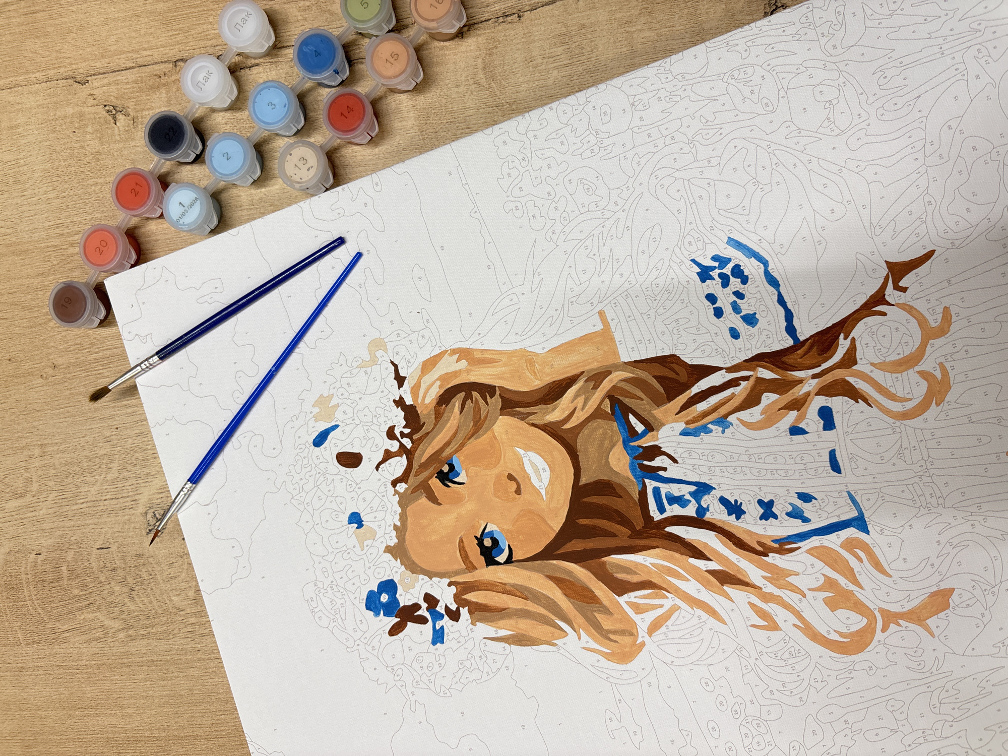 A paint-by-numbers canvas depicting a young girl with blonde hair and blue eyes wearing a floral wreath is laid out on a wooden surface. Two paintbrushes, one blue and one with a brown tip, rest on the canvas. Adjacent to the canvas, a strip of small plastic paint pots, numbered from 1 to 21, is arranged. The paint pots contain various colors of paint. The background is a wooden table. There are no discernible city landmarks or geographical features to indicate a specific location.