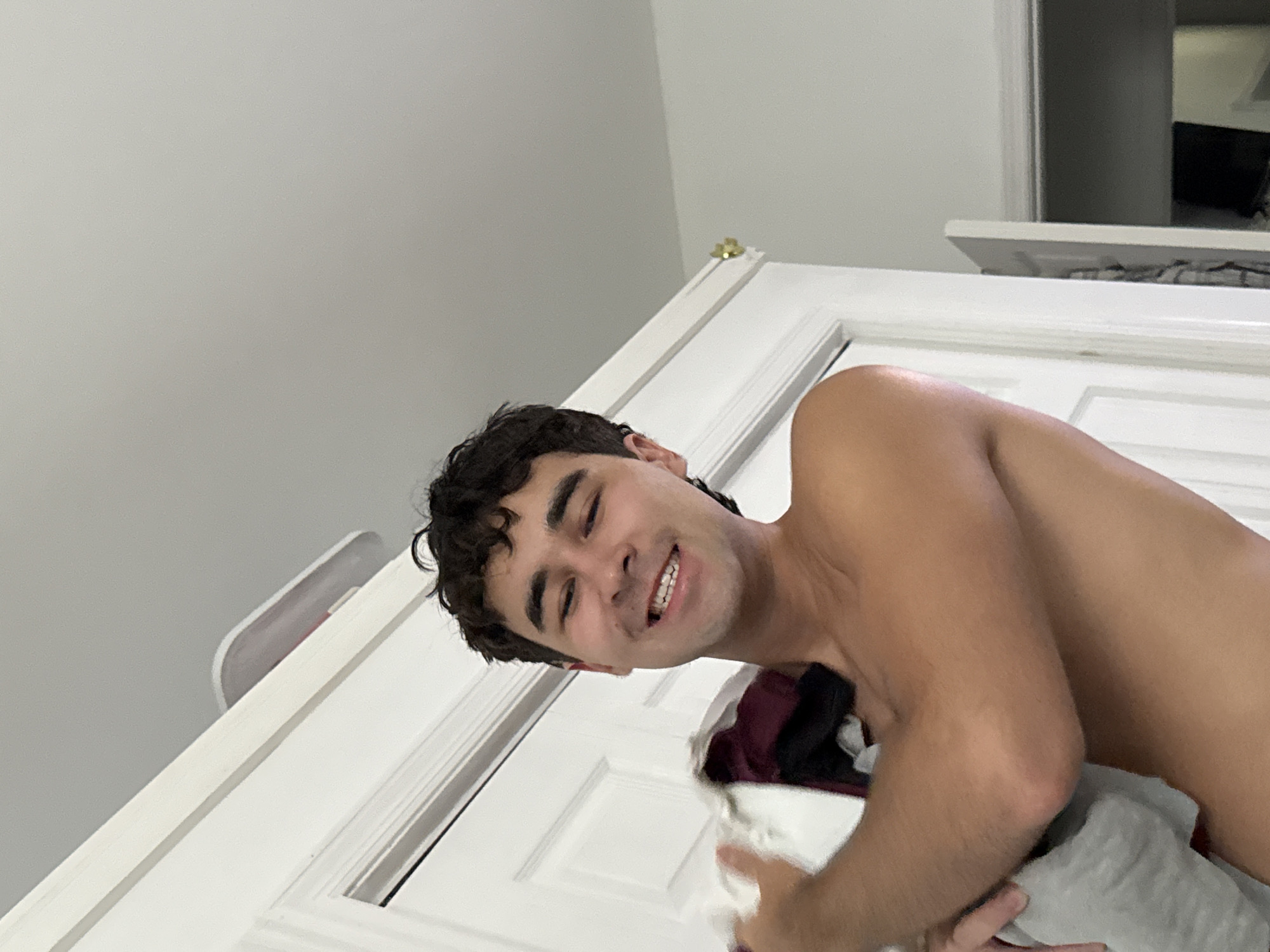 A young man with dark, curly hair smiles and holds a pile of folded laundry to his chest. He is shirtless and his skin is tanned. He appears to be standing in a doorway. The background consists of white doors and a light gray ceiling. On top of the door frame, a white object with a red accent is visible. The scene takes place in a residential interior, likely a home in Bellmore, United States of America.