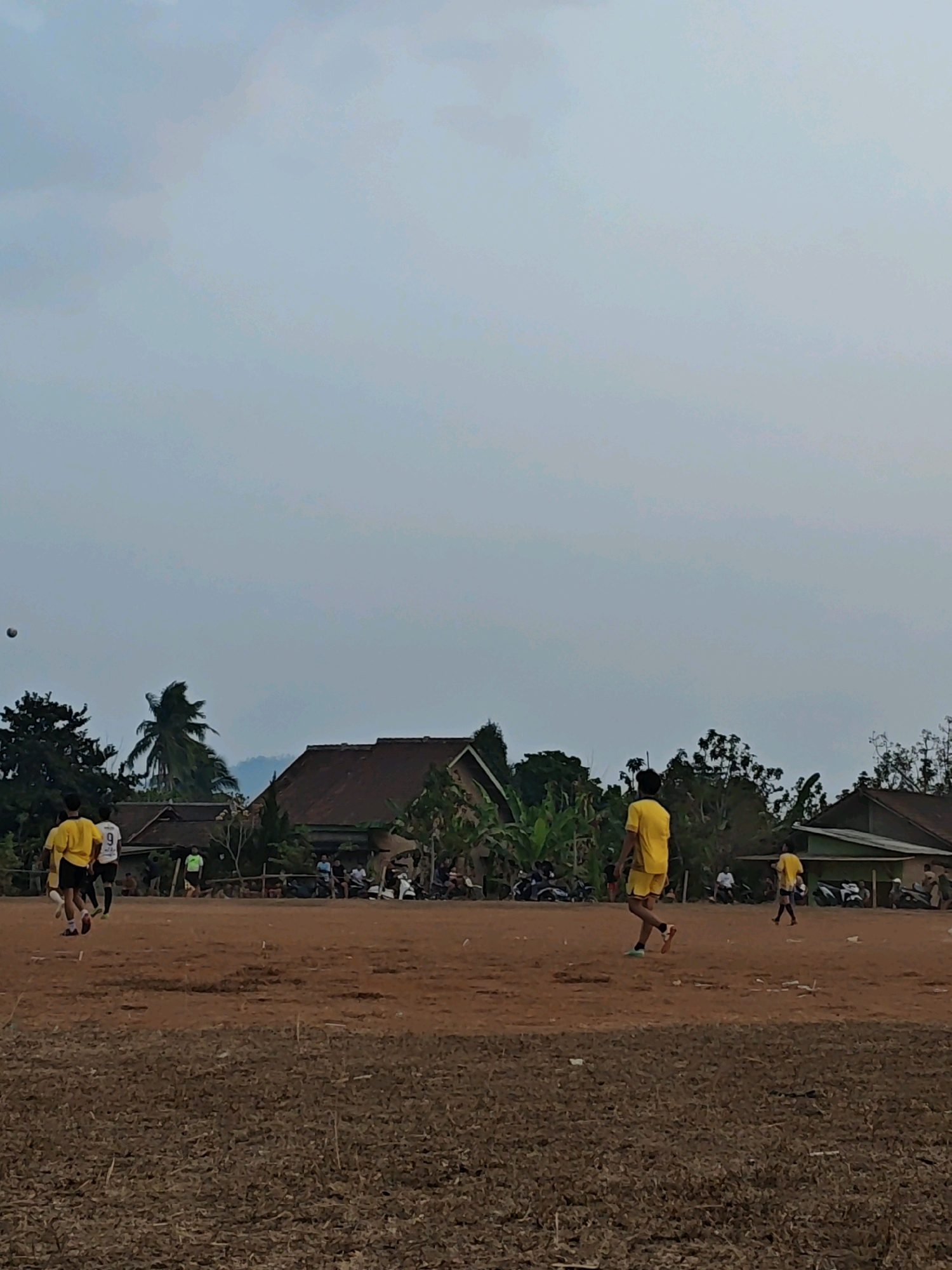 This image depicts a casual soccer match taking place on a dirt field in Karang Sari, Indonesia. The sky is overcast, suggesting either early morning or late afternoon. Several individuals are present, most of whom are wearing yellow soccer uniforms. One person in a yellow shirt and shorts is in motion, appearing to be playing soccer. Another individual in a yellow shirt and white shorts, also in motion, is visible on the left side of the frame. A person in a white shirt with the number 9 on it is also in the foreground. In the background, there are several buildings with reddish-brown roofs, surrounded by lush green trees and palm trees. Some spectators and parked motorcycles are also visible in the background, indicating a local gathering. The overall environment is rural and simple, with the focus on the informal soccer game.