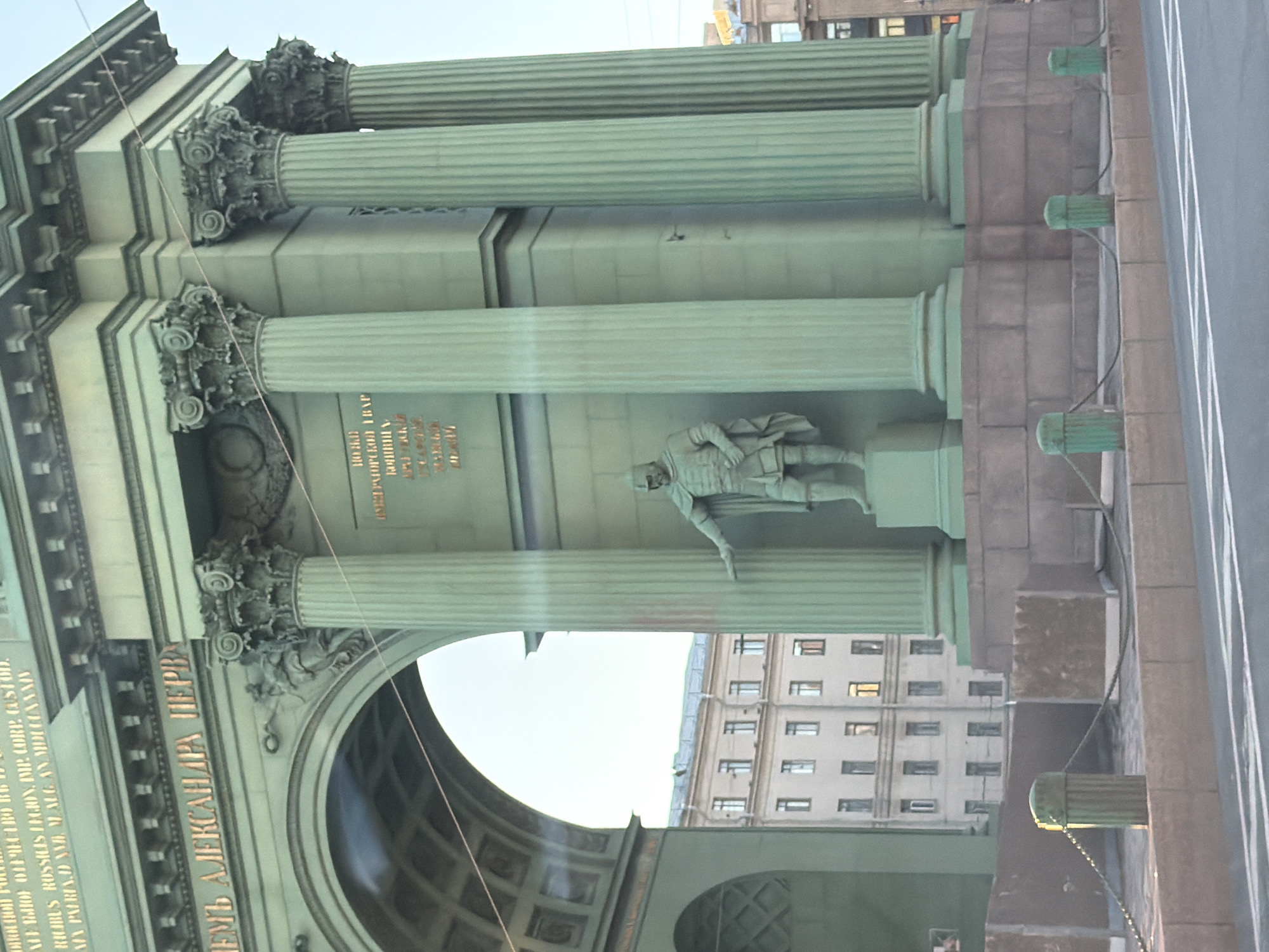 The image shows the Narva Triumphal Arch in Saint Petersburg, Russia. The arch is a pale green color and features three rows of columns with ornate capitals. A sculpture of a soldier is placed on a ledge between the second and third rows of columns. Inscriptions are visible on the arch. A building can be seen through the archway. A gray street surface is also in view.