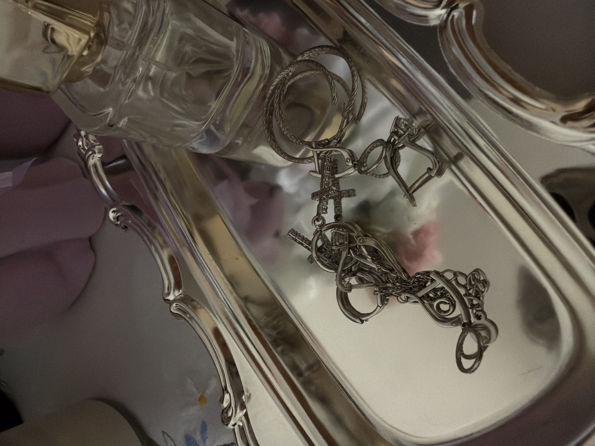 The image depicts a silver-colored tray with ornate edges. Inside the tray is a clear glass bottle, possibly perfume, and various pieces of silver-colored jewelry including hoop earrings, a necklace with a cross pendant, and a chain bracelet. The background shows soft pastel colors, possibly from other objects. There is no indication of Izhevsk, Russia in this image.