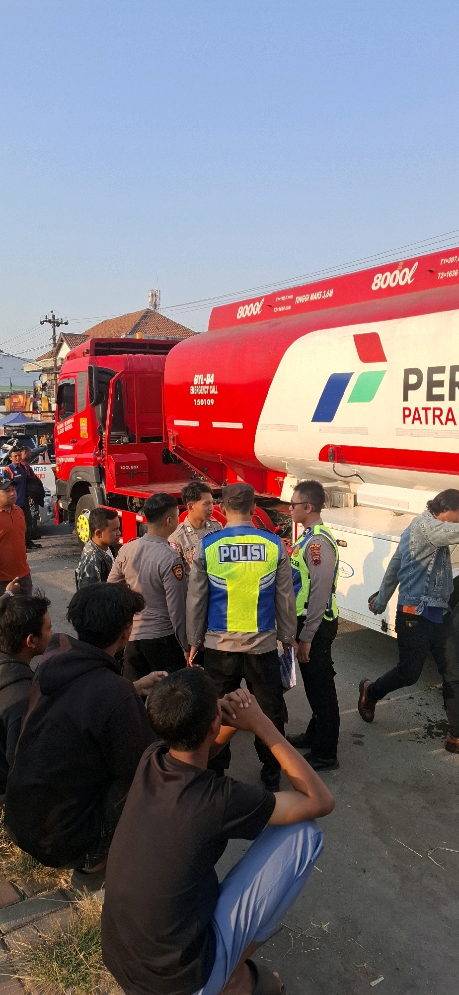 The image depicts a scene in Desa Ngesrep, Indonesia, featuring a red tanker truck with "PERTAMINA" branding on its white tank. Several individuals are gathered around the truck, including at least three men in police uniforms wearing reflective vests with "POLISI" clearly visible. One officer is speaking with a civilian, while others appear to be observing or interacting within the group. The ground is paved with brick and asphalt. The sky is clear and blue, indicating daylight, and there are buildings and utility poles in the background. The overall atmosphere suggests a public gathering or a response to an incident involving the tanker truck.