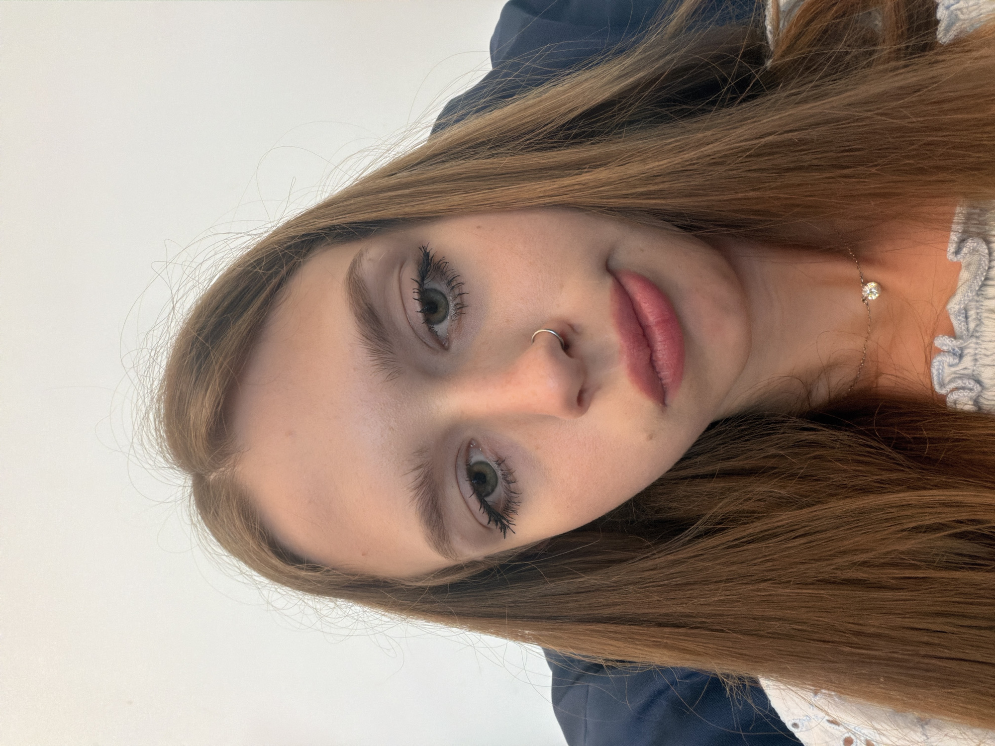 The image is a close-up, over-the-shoulder selfie of a young woman with long, auburn hair. She is looking down and to the side, with a slightly pouty expression. She has green eyes, dark eyelashes, a nose ring, and pink lipstick. She is wearing a white top with eyelet detailing and a silver necklace with a small, round pendant. The background is a plain white wall. The overall impression is one of casual intimacy.