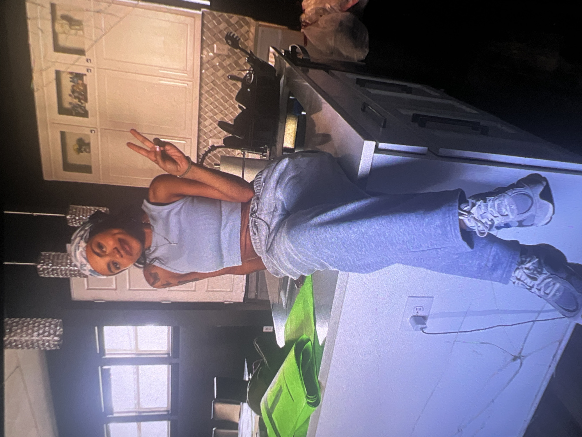 A young woman with a bandana on her head and a light blue crop top is sitting on a kitchen island. She is wearing grey sweatpants and grey sneakers. She is holding up two fingers in a peace sign with her right hand. The kitchen has white cabinets with decorative glass fronts. On the island are two green yoga mats and a black utensil holder filled with kitchen utensils. There is a roll of paper towels to the left of the utensil holder. To the right of the island are plastic bags filled with items. An electrical outlet with a plug in it is visible on the wall below the island. The scene appears to be indoors, in a kitchen. The location is not discernible from the image.