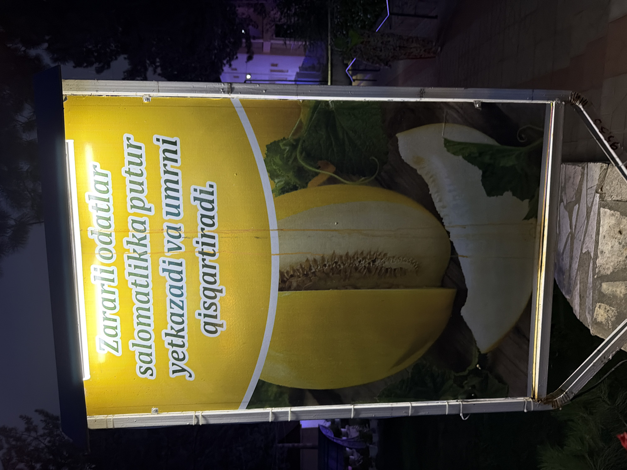 A sign with a yellow background displays text in Uzbek. The text reads, "Zararli odatlar salomatlikka putur yetkazadi va umrni qisqartiradi." Below the text, an image of a halved melon with visible seeds and a slice of melon is depicted. The sign is illuminated by a bright light above it. In the background, a person is visible on the right side of the image, wearing camouflage clothing. The scene appears to be outdoors at night, with trees and buildings visible in the background, suggesting a location in Khumsan, Uzbekistan.