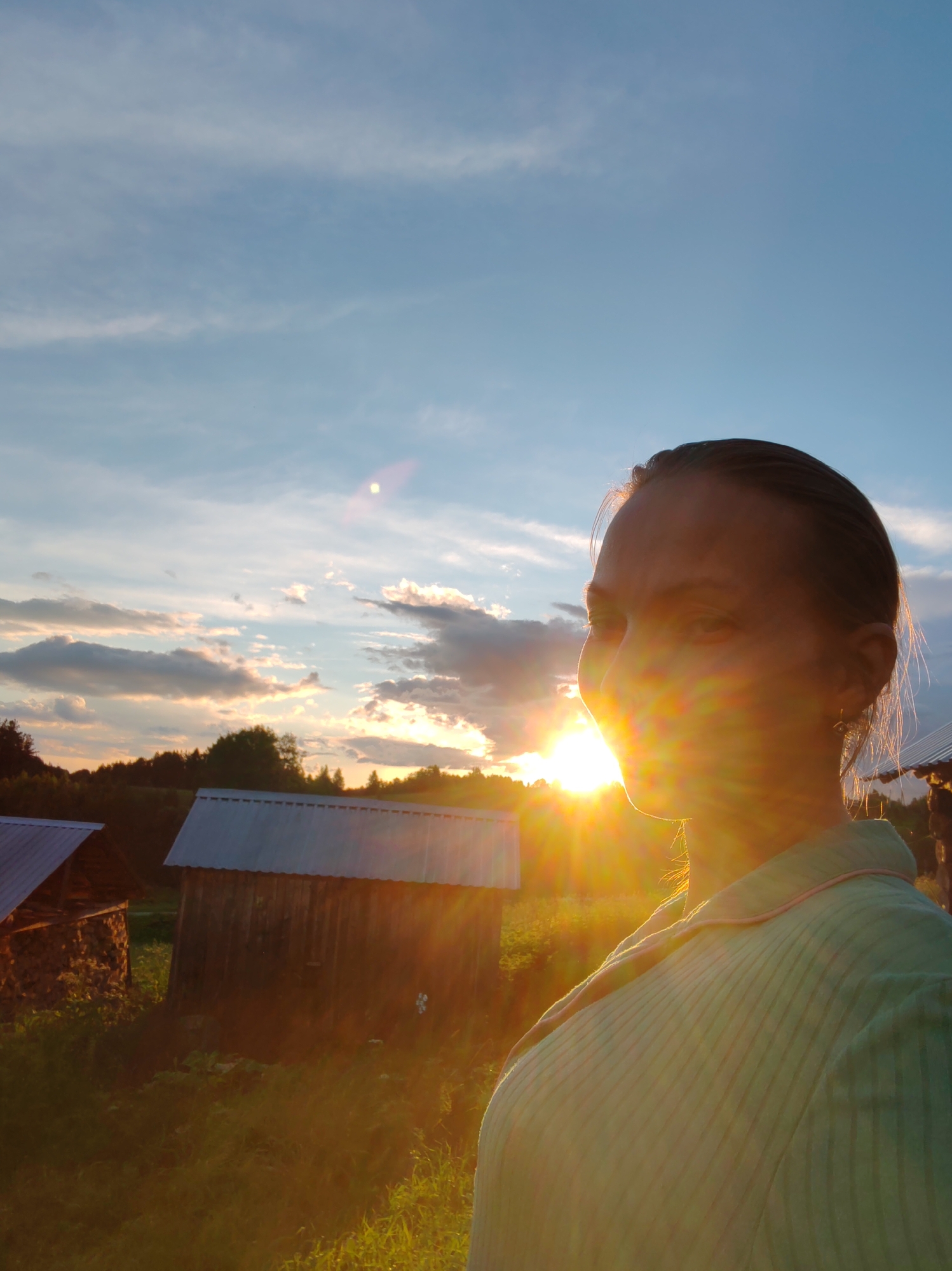 The image captures a young woman in profile against a backdrop of a rural sunset, likely in the village of Chukhlem, Russia. The sun, positioned low in the sky, casts a strong golden light that flares across the frame, obscuring details of the woman's face and creating lens flare effects. She is wearing a light-colored, collared shirt with vertical ribbed texture and a pink trim along the collar. Her hair is pulled back, revealing her ear with a small earring.
In the background, two rustic wooden structures with metal roofs are visible. The structure on the left appears to be stacked with firewood or logs, while the structure on the right is a more traditional outbuilding. A dense line of trees forms the horizon, silhouetted against the colorful sky, which is a gradient of blues and oranges with scattered clouds. The foreground is filled with tall, sunlit grass, suggesting a natural, uncultivated environment. The overall impression is one of a serene, idyllic rural setting bathed in the warm glow of the setting sun.