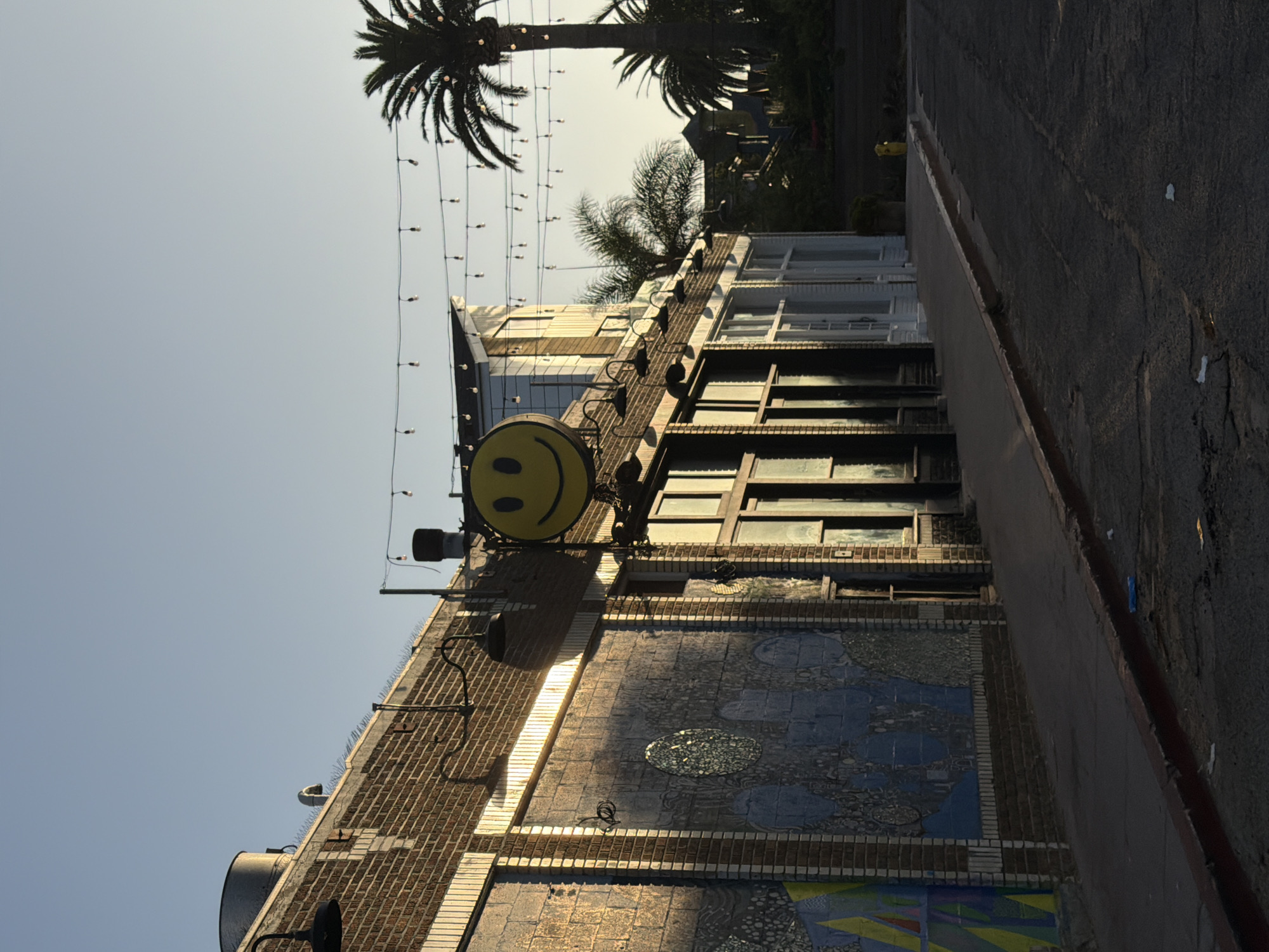 The image captures a street scene in Los Angeles, California, at dusk. The setting sun casts long shadows across the asphalt road and illuminates the brick facade of a building on the left. A large, yellow smiley face sign hangs above the building's entrance, adding a whimsical touch to the urban landscape. String lights are suspended across the street, casting a warm glow as twilight descends. Palm trees are visible in the background, their fronds silhouetted against the pale sky, further emphasizing the Southern California setting. The building features large windows, some reflecting the warm light of the setting sun. The overall impression is one of a quiet, late afternoon in a city known for its vibrant street art and relaxed atmosphere.