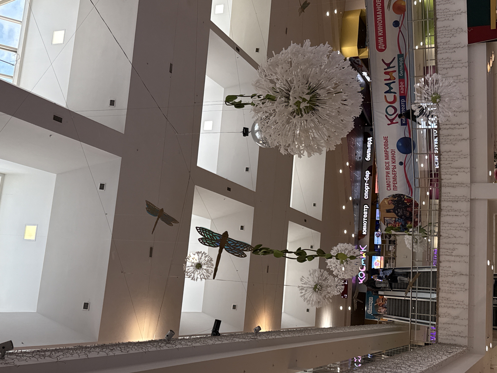 This image depicts the interior of a multi-story commercial building, likely a shopping mall or entertainment complex, viewed from a low angle looking upwards. The setting features high ceilings with stark white and light grey walls, punctuated by rectangular windows or skylights through which a bright blue sky with some clouds is visible, indicating daytime.
Numerous decorative elements are suspended from the ceiling. These include large, spherical white ornaments resembling dandelions, and several stylized wooden dragonflies, some with blue and green patterns on their wings. A silver disco ball is also visible, hanging near one of the white spheres. The architecture incorporates multiple levels, with railings adorned with string lights and spotlights. Rectangular light fixtures are set into the ceiling panels, and ventilation grilles are visible on the walls.
Prominently displayed in the background are large banners and signs for an entertainment center named "КОСМИК" (Kosmik). The text on these banners advertises "ДНИ КИНОМАНОВ" (Days of Movie Lovers) and urges patrons to "СМОТРИ ВСЕ МИРОВЫЕ ПРЕМЬЕРЫ КИНО!" (Watch all world cinema premieres!). Services offered at Kosmik include "кинотеатр" (cinema), "кафе" (cafe), "бильярд" (billiards), and a "спорт-бар" (sports bar). Another sign partially reads "ЕСЛИ ЭБУВЬ, 10". A few indistinct individuals can be seen near escalators on a lower level, suggesting general public activity within the venue.