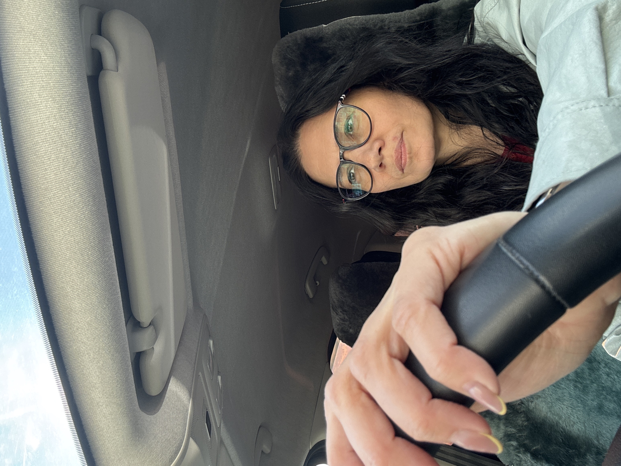 This image captures a woman inside a car, reportedly in Chaikovsky, Russia, from a low-angle perspective looking upwards. The woman, likely the driver, has long, dark, wavy hair and wears eyeglasses with round frames. She is dressed in a light-colored jacket over a dark red top, and her expression is neutral to slightly contemplative. Her right hand is prominently featured in the foreground, gripping the black steering wheel, with her fingernails showcasing a French manicure style with yellow tips. The car's interior is visible, including a light gray headliner and a sun visor that is folded up. Bright sunlight streams through the windshield, indicating it is daytime. There is no visible text in the image.