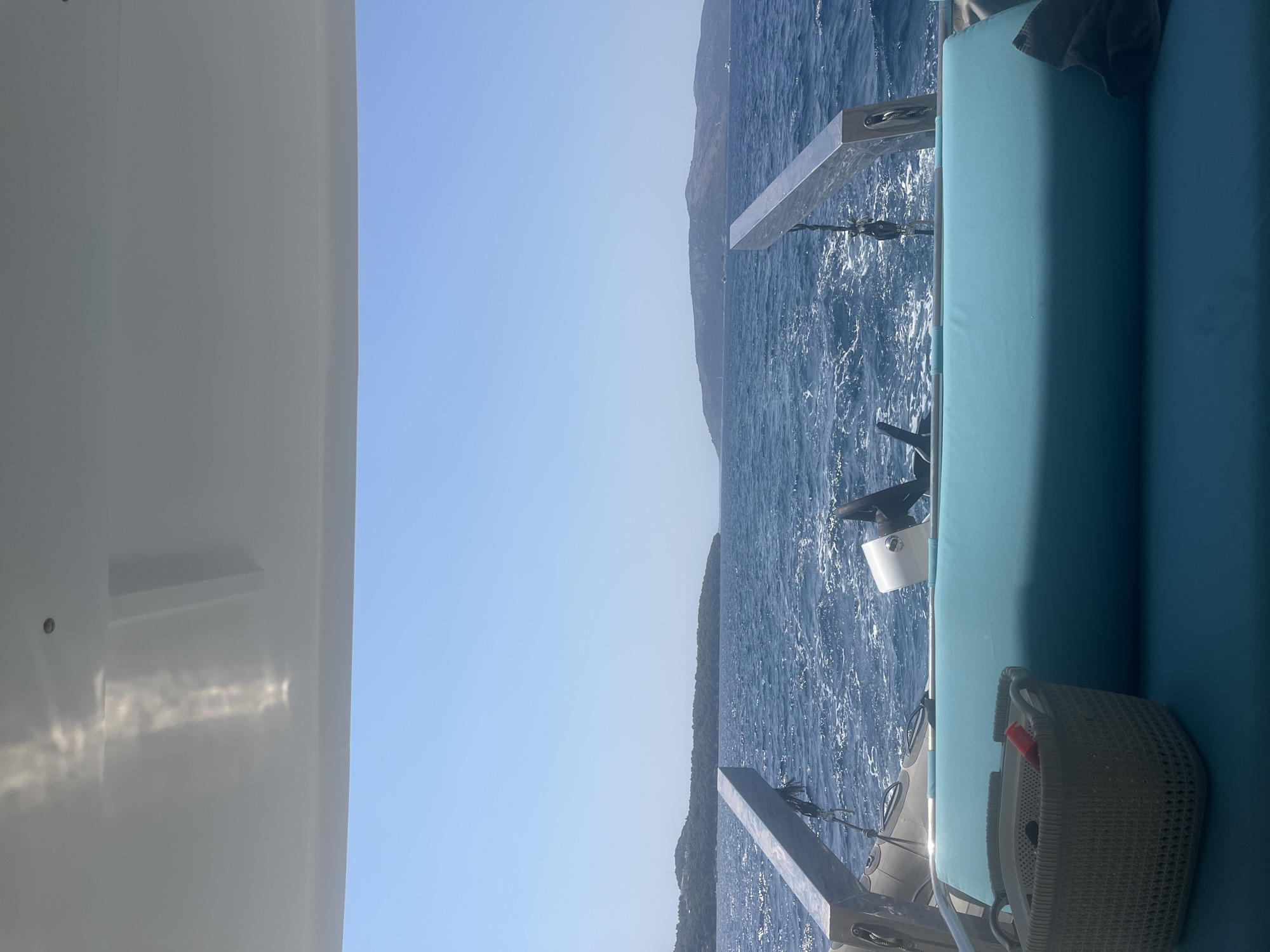 The image captures a serene view from a boat navigating the Adriatic Sea, likely near Okrug Donji, Croatia. The foreground features a turquoise cushioned seat with a woven grey basket containing a red object. To the left, a silver railing extends outward, with a grey inflatable boat partially visible beneath it. The middle ground is dominated by the deep blue, choppy waters of the sea, with a white wake trailing behind the boat. In the distance, lush green islands rise from the water under a clear, bright blue sky. The sun casts reflections on the underside of the boat's canopy, creating bright streaks of light. The scene evokes a sense of peaceful travel and vacation.