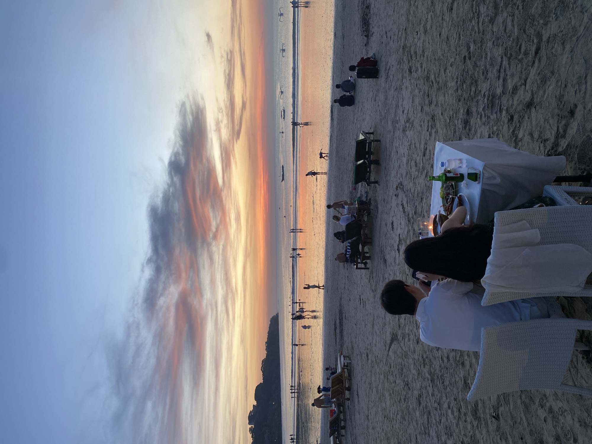 The image depicts a beach scene at sunset in Kuta Selatan, Indonesia. The sky is a mix of blue, orange, and pink hues. Numerous individuals are present on the beach, some walking along the shoreline and others seated. In the foreground, a couple is sitting at a dining table set up on the sand, complete with a white tablecloth, drinks, and dinnerware. The image captures a romantic setting at the beach.