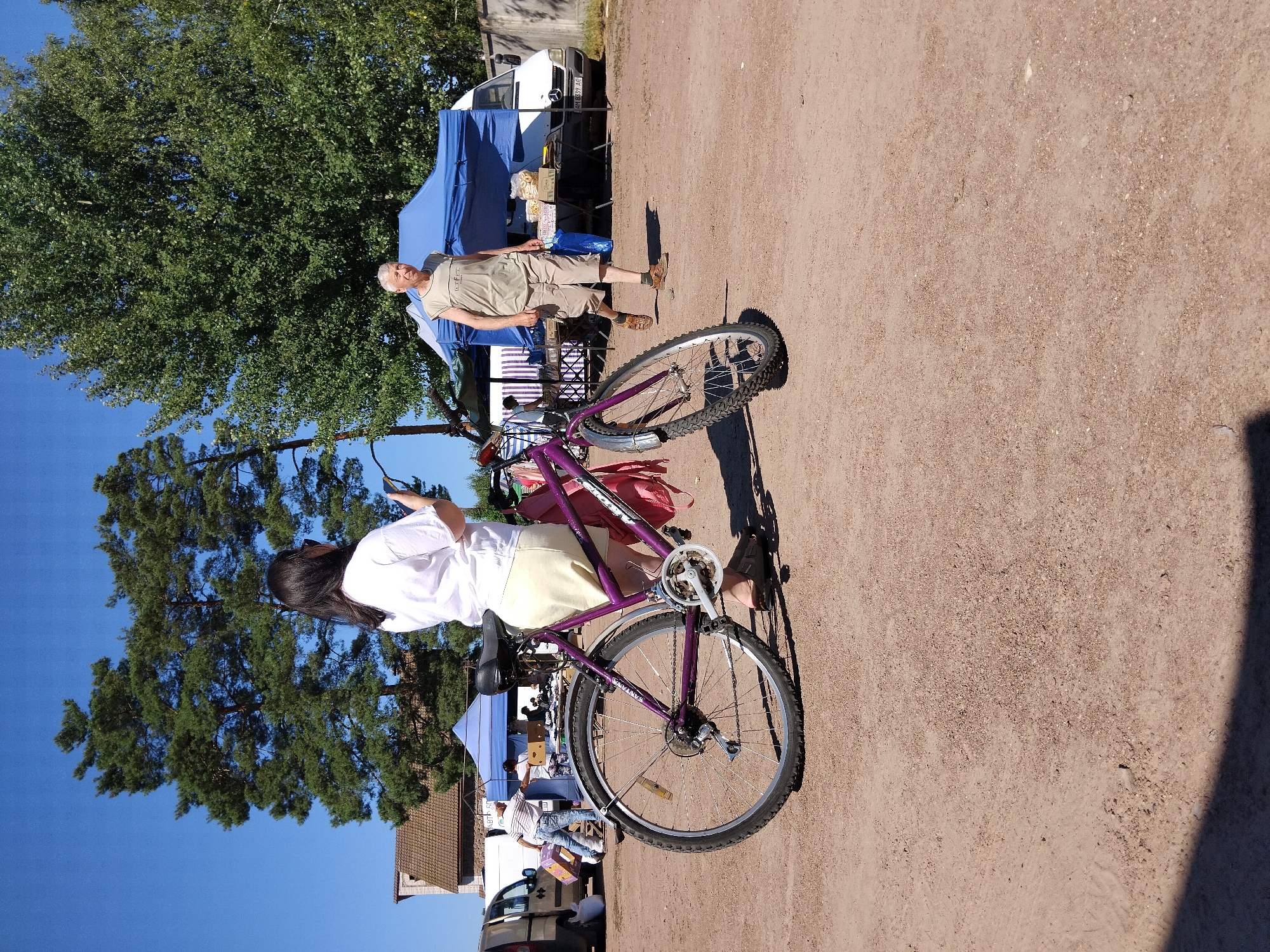 A medium shot captures a woman from behind, holding a smartphone, standing next to a purple bicycle. She is wearing a white t-shirt and a light-colored skirt. The bicycle has a pink bag attached to its rear. In the background, a man with gray hair and wearing a tan sleeveless shirt and shorts walks away from a market stall with blue tents. Behind him, a white van is parked. The scene is set outdoors on a dirt surface under a clear blue sky, with large trees visible in the background. The location is a market in Irshansk, Ukraine.