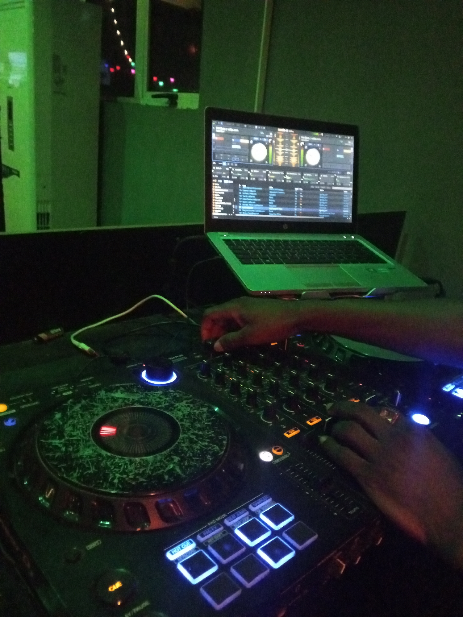 In a dimly lit environment in Lokoja, Nigeria, a DJ operates a digital DJ controller connected to a laptop. The DJ's hands are visible manipulating the mixer's controls. The laptop displays DJ software. The controller has illuminated buttons and a jog wheel. The environment is illuminated with green and blue lights.