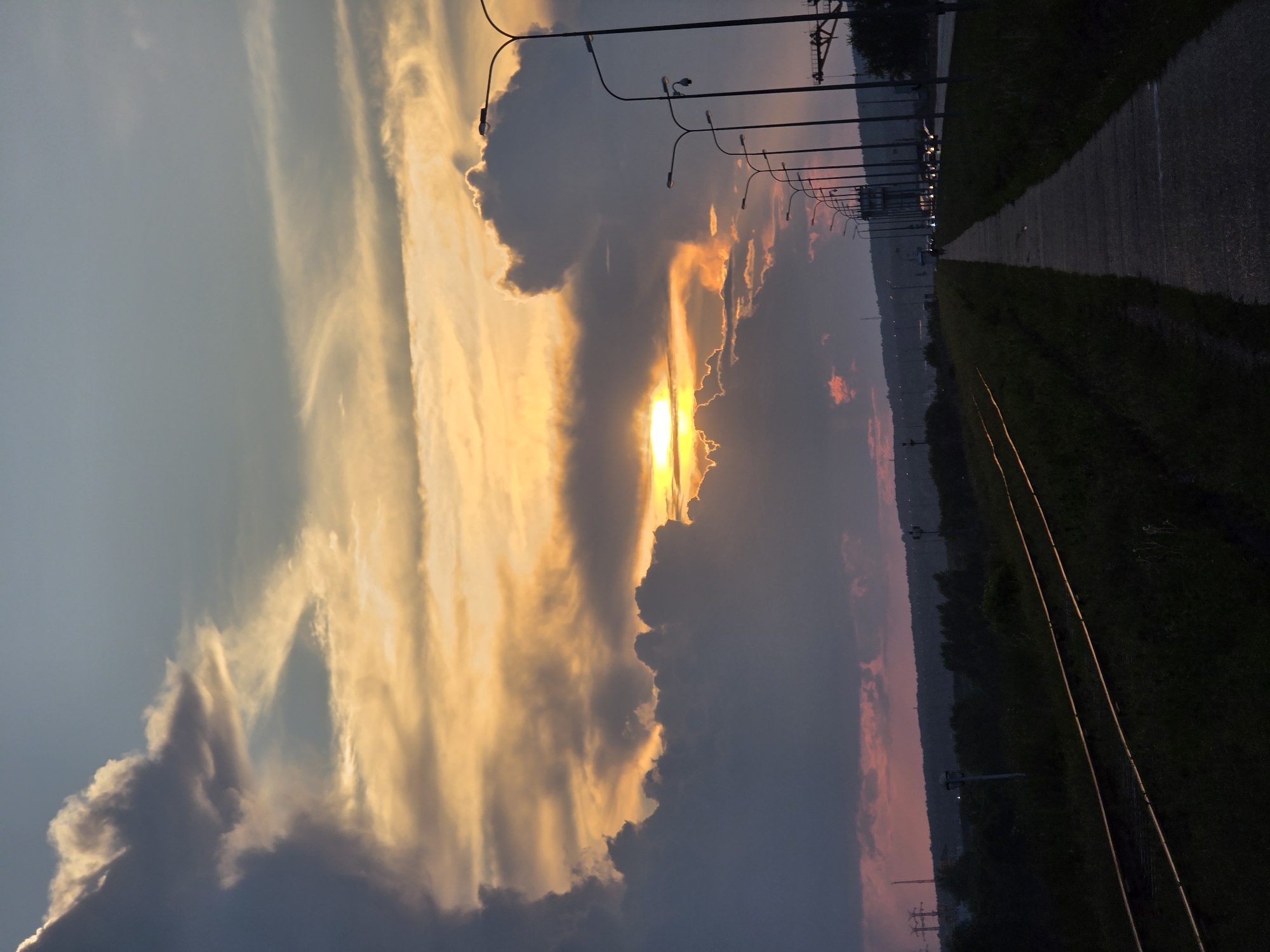 The image captures a dramatic sunset over Chaikovsky, Russia. The sky is filled with a riot of colors, from deep blues and grays in the lower clouds to vibrant oranges and yellows in the upper layers where the sun is partially obscured. Jagged, dark clouds dominate the left side of the frame, contrasting with the soft, luminous glow emanating from the setting sun.
In the foreground, a disused railway track runs parallel to a paved footpath, both disappearing into the darkening landscape. The tracks are edged by overgrown grass and vegetation. To the right of the footpath, a line of modern streetlights stretches along a road, some of which are illuminated, casting a faint light on the passing vehicles visible in the distance. The overall impression is one of the natural beauty of the sunset juxtaposed with the man-made infrastructure of the city.