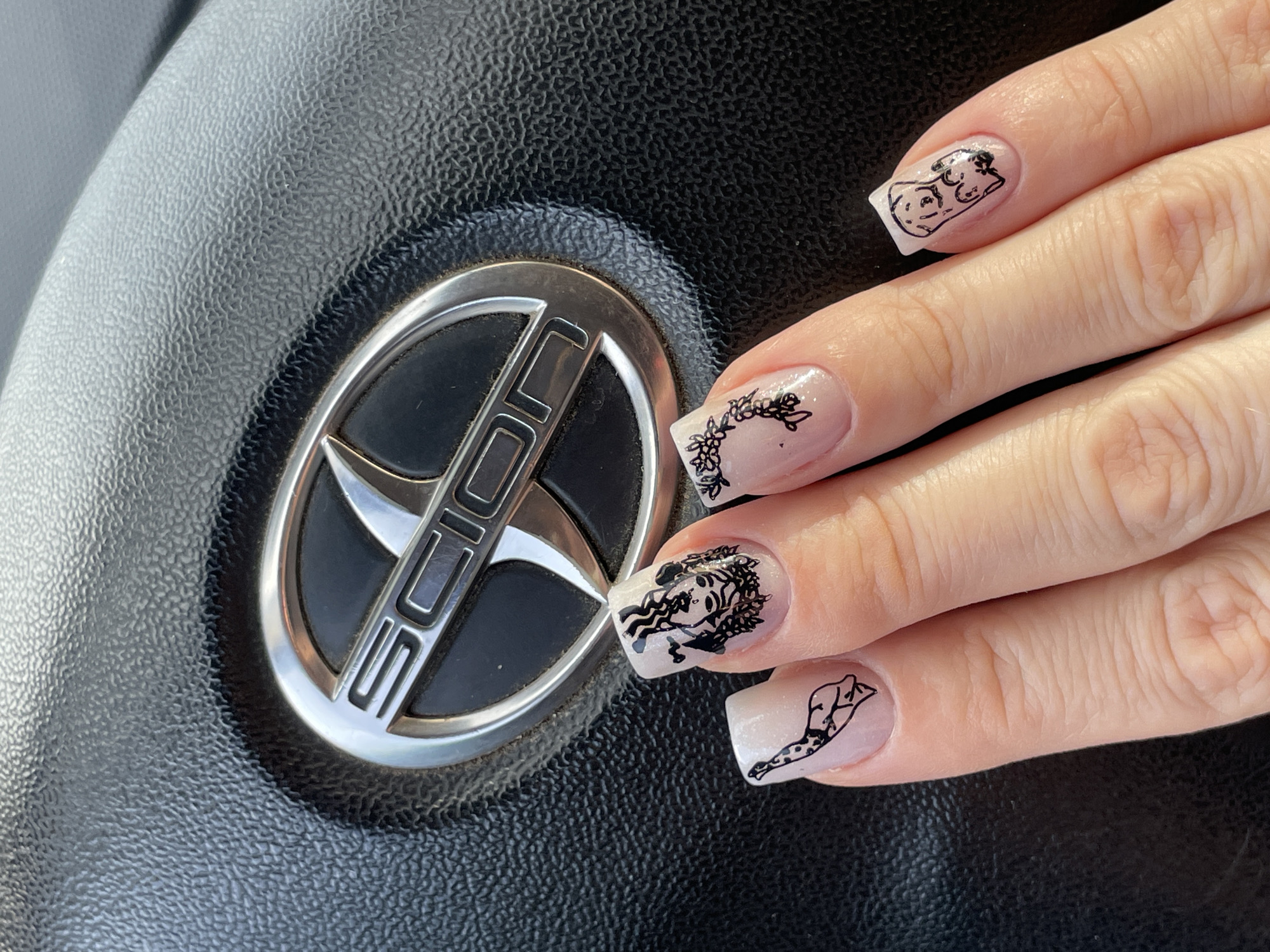 A hand with square-shaped, light-colored fingernails adorned with black line art is held in front of a black, textured steering wheel with a silver and black "SCION" logo in the center. The nails feature various designs: the thumb has a Dalmatian dog illustration, the index finger has a close-up of a face with flowing hair and floral elements, the middle finger has a delicate floral wreath, and the ring finger has an illustration of two puppies. The background suggests an outdoor setting with natural light casting shadows. The location is unspecified, and there are no indications of Aktau, Kazakhstan.