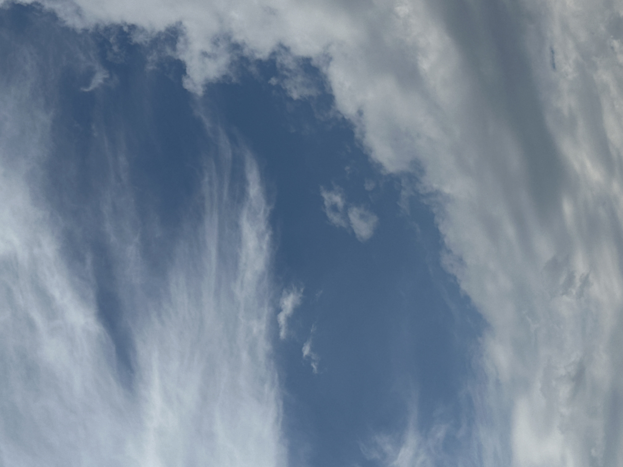 The image is a close-up, full-frame shot of a sky with clouds. The dominant color is blue, indicative of a clear sky, interspersed with various types of clouds. Wispy cirrus clouds stretch across the upper portion of the frame, appearing as thin, feathery streaks. Below these, larger, puffier cumulus clouds are visible, some appearing dense and white, while others are darker gray, suggesting they may be carrying moisture or are in shadow. The clouds are distributed unevenly, with a significant mass of white and gray clouds occupying the right and bottom edges of the image, while a clearer blue expanse dominates the center and left. The lighting suggests daylight, with the sun likely positioned above and to the right of the frame, casting shadows on the denser clouds. There are no visible signs of human activity, structures, or landmarks that would indicate a specific location within the city of Makiv, Ukraine, or any other urban environment. The image is a straightforward depiction of a sky on a day with mixed cloud cover.