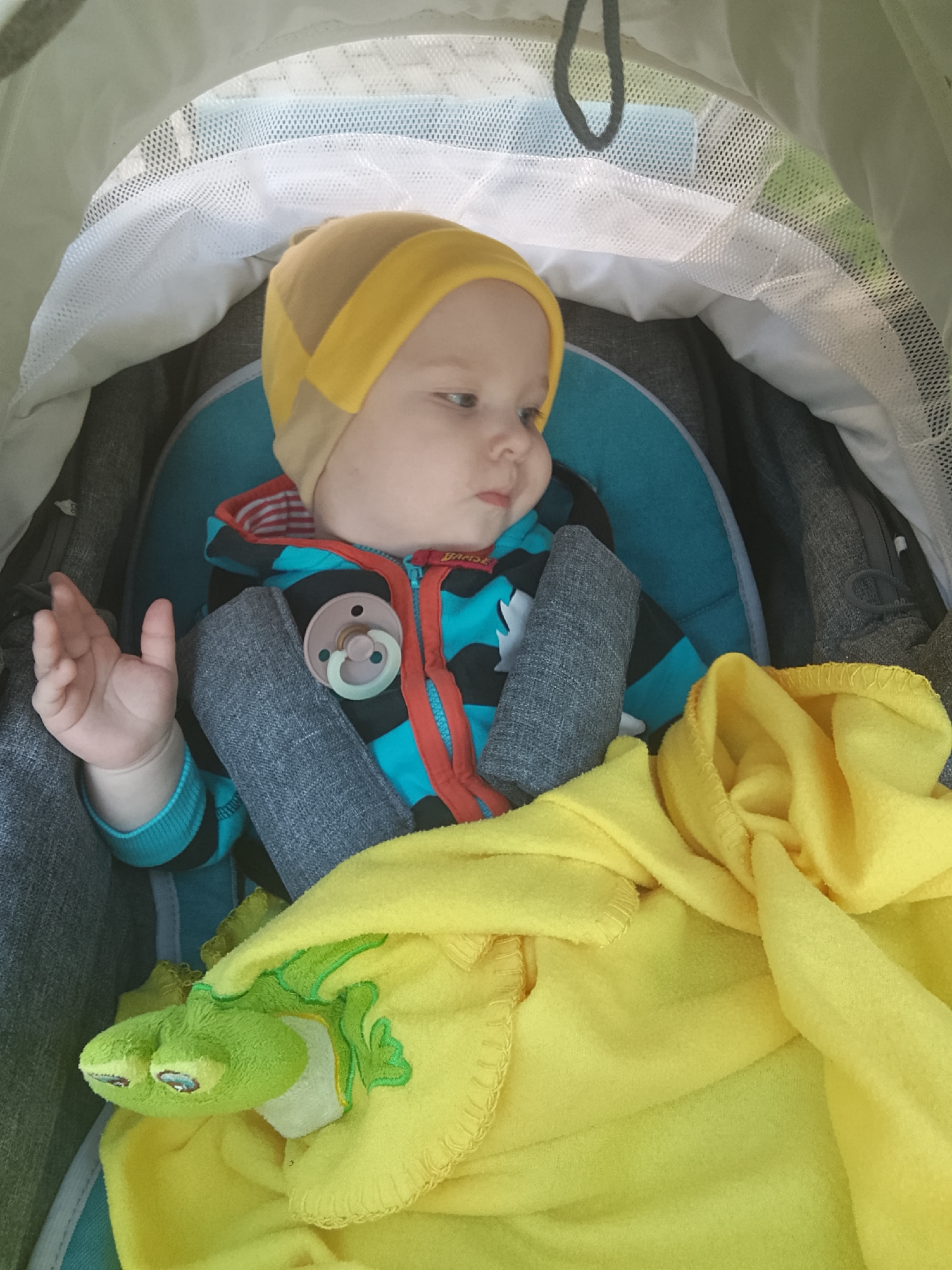 A close-up, eye-level shot shows a baby boy sitting in a stroller. The baby is wearing a yellow and brown striped beanie, a blue and black striped long-sleeved shirt with red trim, and a pacifier with a pink ring. He is looking to the right, with his left hand raised. The baby is covered with a yellow blanket, which has a green stuffed frog peeking out from under it. The stroller has a grey frame and a blue padded seat. The background is blurred but appears to be an outdoor setting with some greenery. The lighting is bright, suggesting it is daytime. The image was taken in Minsk, Belarus.