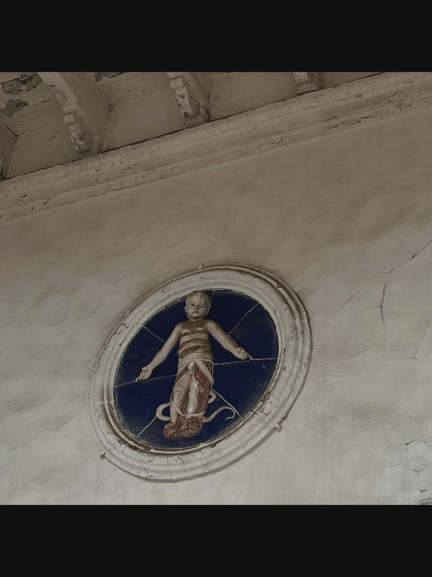 In Bielawska Wieś, Poland, a faded oval fresco is visible on a weathered, light-colored wall. The fresco depicts a child-like figure with outstretched arms, wrapped in a reddish-brown and white cloth. The background of the fresco is a dark blue color, divided into segments by thin white lines. Above the fresco, the ceiling is adorned with decorative beams and trim, showing signs of age and peeling paint. The wall itself exhibits numerous cracks, indicating wear and tear.