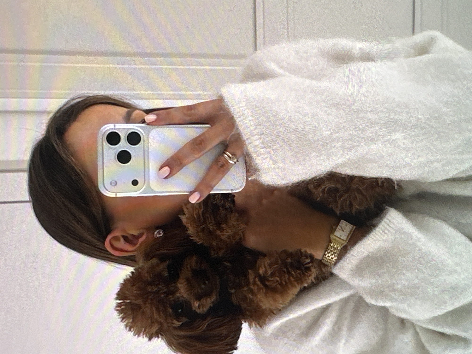 A person is taking a selfie in a mirror, holding a brown poodle dog close to their body. The person is wearing a white sweater, a gold watch, and a ring. Their fingernails are painted a light pink color. The cell phone they are using to take the selfie has a clear case and three camera lenses. The background appears to be a white wall with horizontal lines. There is no evidence to suggest this image was taken in Yerevan, Armenia.