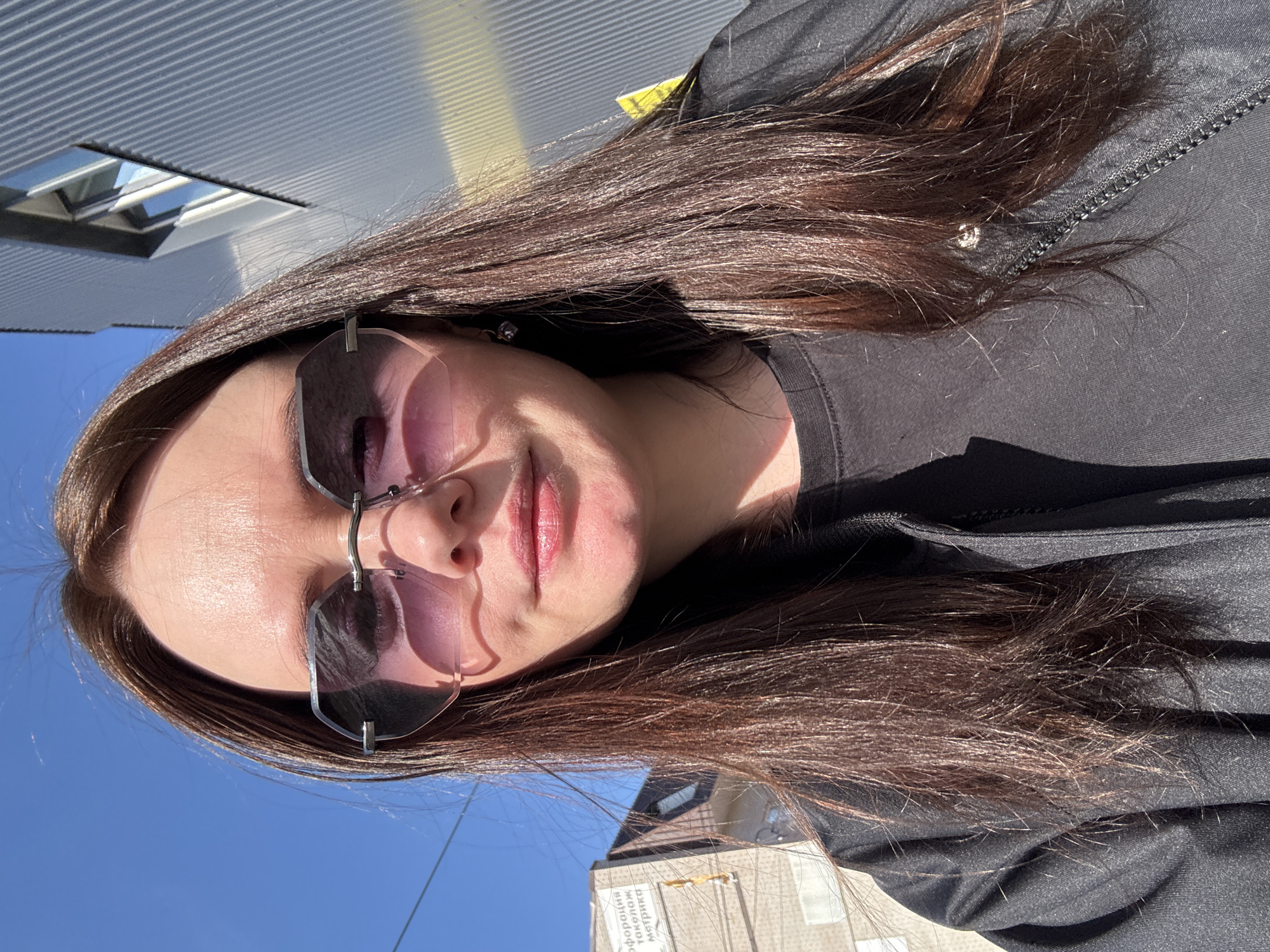 A female individual is centrally positioned outdoors under a clear blue sky, illuminated by strong sunlight from the upper right. She has long, dark brown hair, wears rimless hexagonal sunglasses with purple-tinted lenses, a black t-shirt, and a black zippered jacket. A dark earring is visible on her right ear, and she exhibits a slight smile. The background on the left features an older, light-colored building (likely brick or stucco) with exposed wiring. Affixed to this building is a sign with Cyrillic text reading "ИНФОРМАЦИЯ" (INFORMATION), "токелаж" (rigging/tackle), and "метрика" (metrics/metrology). To the right, a modern building with grey corrugated metal siding is visible, featuring a recessed window with a diagonal bottom edge that reflects the sky. A faint yellow stripe or reflection appears on the lower portion of the modern building. The Cyrillic sign text, particularly "токелаж" and "метрика," identifies the location as the city of Chaikovsky, Perm Krai, Russia, likely within a commercial district.