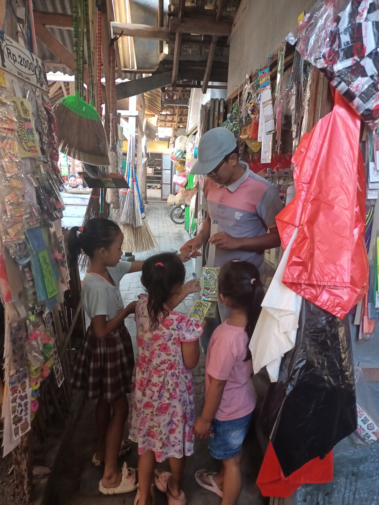 In Badas, Indonesia, a vendor wearing a grey baseball cap and a pink and grey polo shirt interacts with three young girls in a market stall. The vendor is handing something to one of the girls. The stall is lined with various items, including brooms, bags, and small packaged goods. In the background, a motorcycle is visible. The overall scene depicts a lively marketplace.
