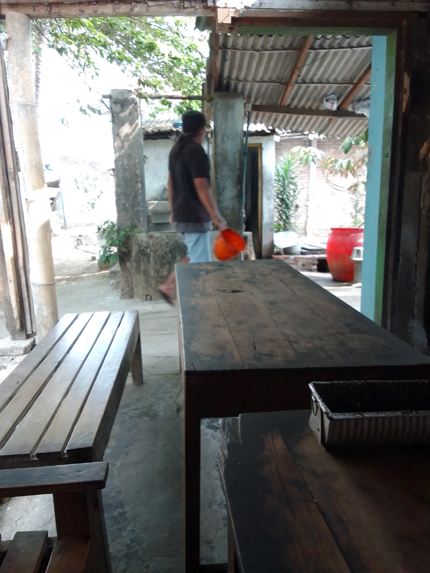 This image depicts an outdoor scene in Trenggalek, Indonesia, likely a food stall or restaurant. In the foreground, a wooden bench is visible, followed by a dark wooden table with a metal tray on it. To the right, a bright red barrel is prominent in the background. A person wearing a dark shirt and light blue shorts is walking away from the camera, carrying an orange bucket. The environment includes a corrugated metal roof, concrete floor, and vegetation. The overall impression is of a rustic, casual setting.