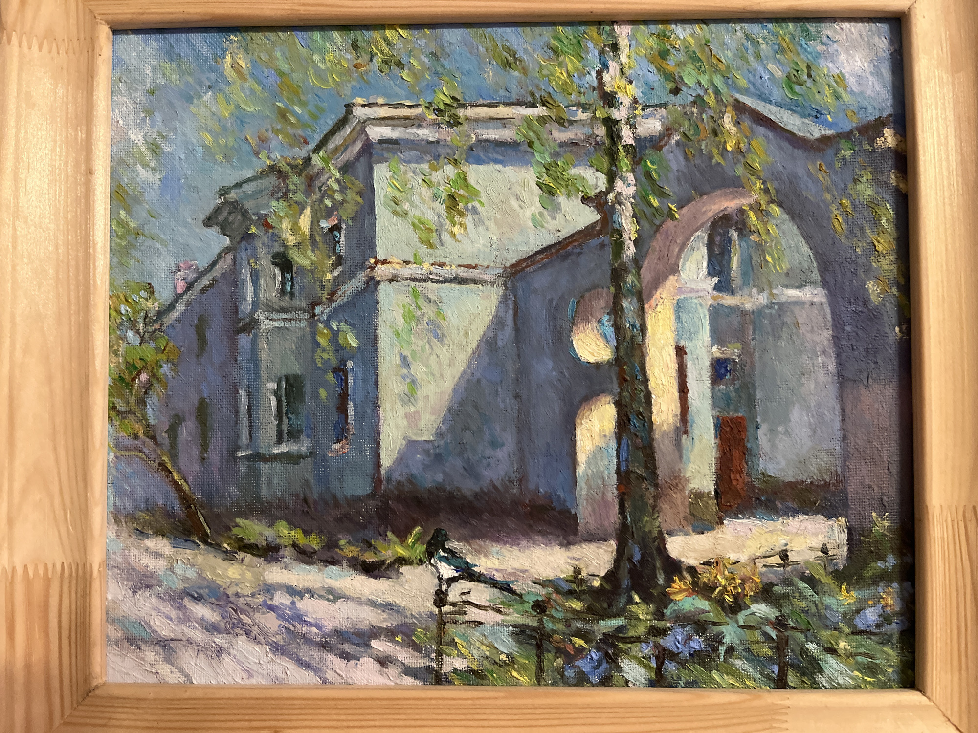 The image is a painting in a wooden frame, depicting a building with a large arched entrance. The building is painted in muted tones of blue and gray, with a white roofline. Sunlight casts strong shadows across the facade. In front of the building, a tree with a textured bark stands in the foreground, its leaves painted in dabs of green and yellow, suggesting dappled sunlight. A small bird, possibly a magpie, is perched on a fence in the lower left of the painting. The ground in front of the building is rendered with loose brushstrokes in shades of white, purple, and yellow, indicating a path or ground cover. The sky above is a bright blue with streaks of white. The style is impressionistic, with visible brushstrokes and a focus on light and color. This scene is identified as the building of the former church of St. John the Baptist in Starye Politsy, Russia, which now serves as a cultural center.