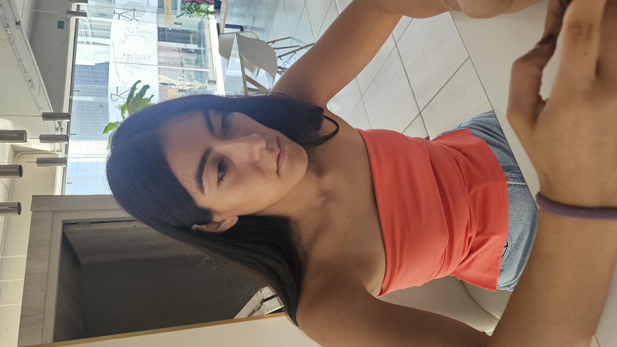 A young woman with dark hair and an orange top is sitting at a table. She is wearing jeans. The location appears to be a modern indoor space, potentially a cafe or office in Puebla, Mexico, given the architectural style visible through the window. There are white chairs in the background. The woman is looking towards the camera.
