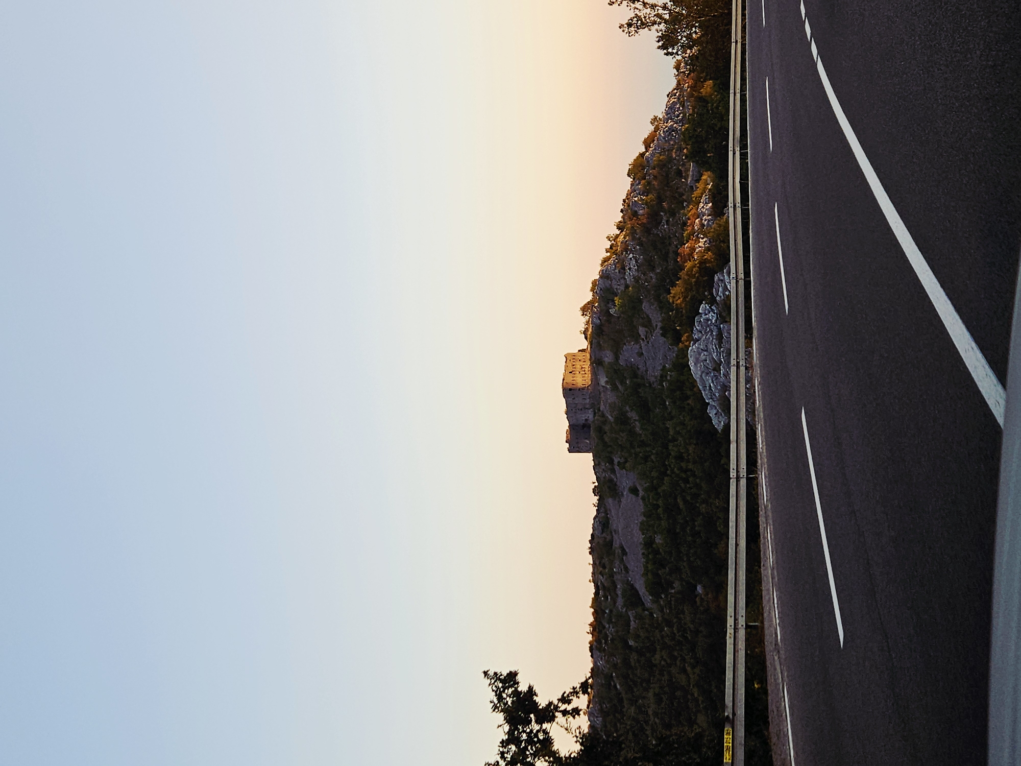 The image shows a road in the foreground with white lane markings, leading towards a hilly landscape under a clear, pale blue sky. A metal guardrail separates the road from the embankment. On top of a prominent hill, a stone structure, possibly a fort or an old building, is visible. The hill is covered with green vegetation and scattered rocks. The setting sun casts a warm glow on the landscape, creating a soft gradient of orange and pink hues in the sky near the horizon. A few trees are present on the left and right sides of the frame, framing the scene. The overall impression is one of a scenic, possibly rural, location at dusk or dawn.