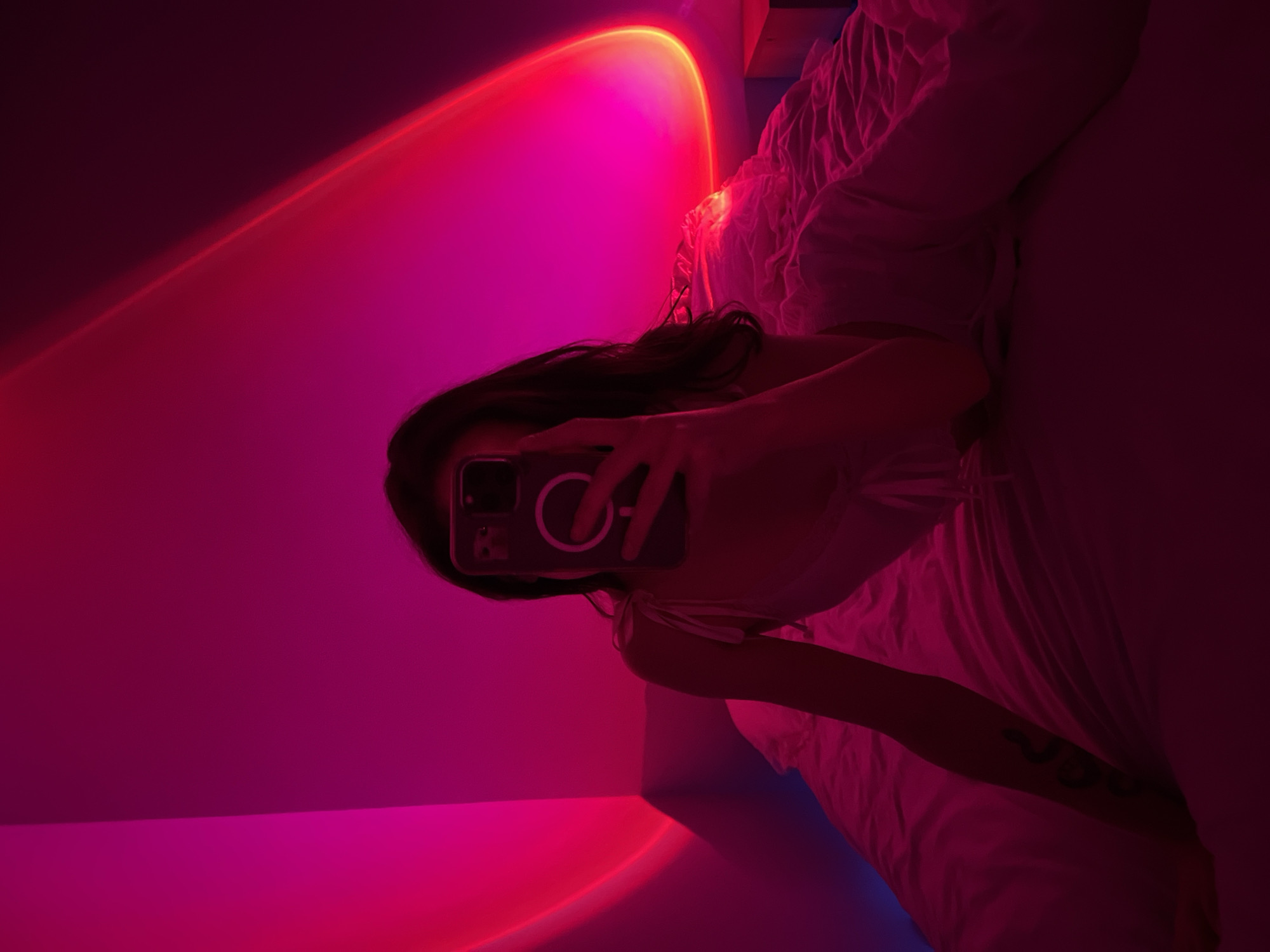 The image depicts a person holding a smartphone, taking a selfie in a bedroom. The room is illuminated by a pink and red sunset lamp, casting a gradient of color across the walls and a bed covered with white sheets. The person is partially visible, with their hair dark and long, and they appear to be wearing light-colored clothing. A tattoo is visible on the person's left forearm. The overall mood of the image is intimate and atmospheric, enhanced by the dramatic lighting. There is no information in the image to suggest a location in Ternopil, Ukraine.