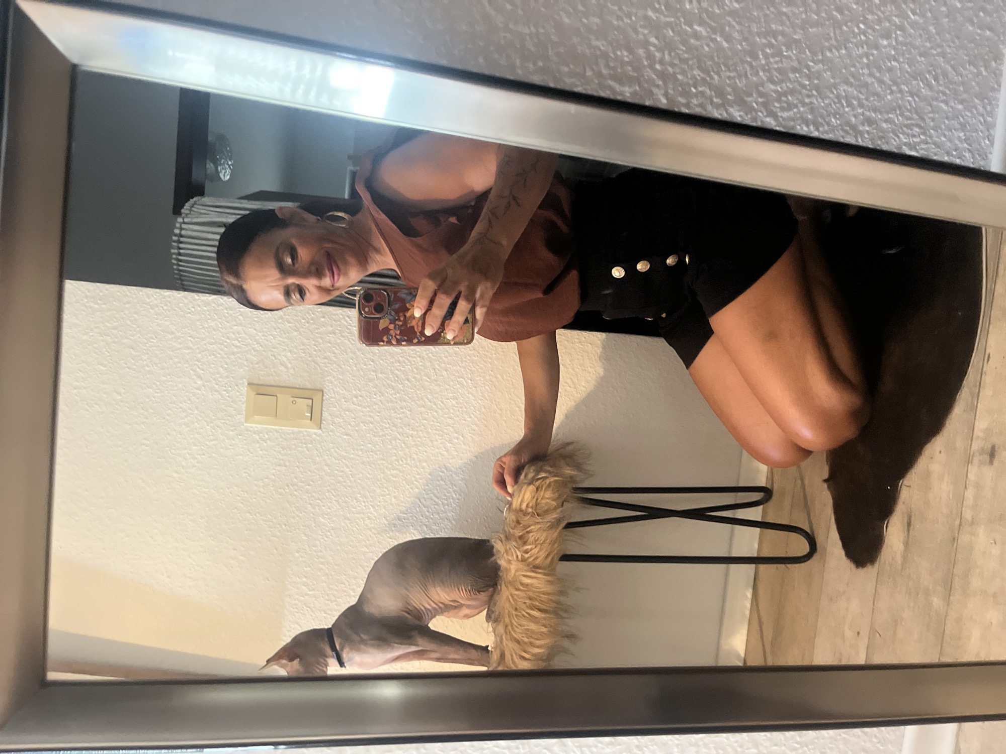 A woman is taking a mirror selfie while kneeling. She is wearing a brown ruffled top and black shorts with gold buttons. She has a phone with a floral case in her right hand. Next to her, a hairless cat is sitting on a fluffy stool. The scene appears to be indoors, possibly in an apartment or house. There is a light switch on the wall behind the cat. The floor is made of light-colored wood planks, and there is a dark rug underneath the stool. The mirror frame is dark and metallic. The image does not provide any information to determine the location in the city of Konz, Germany.