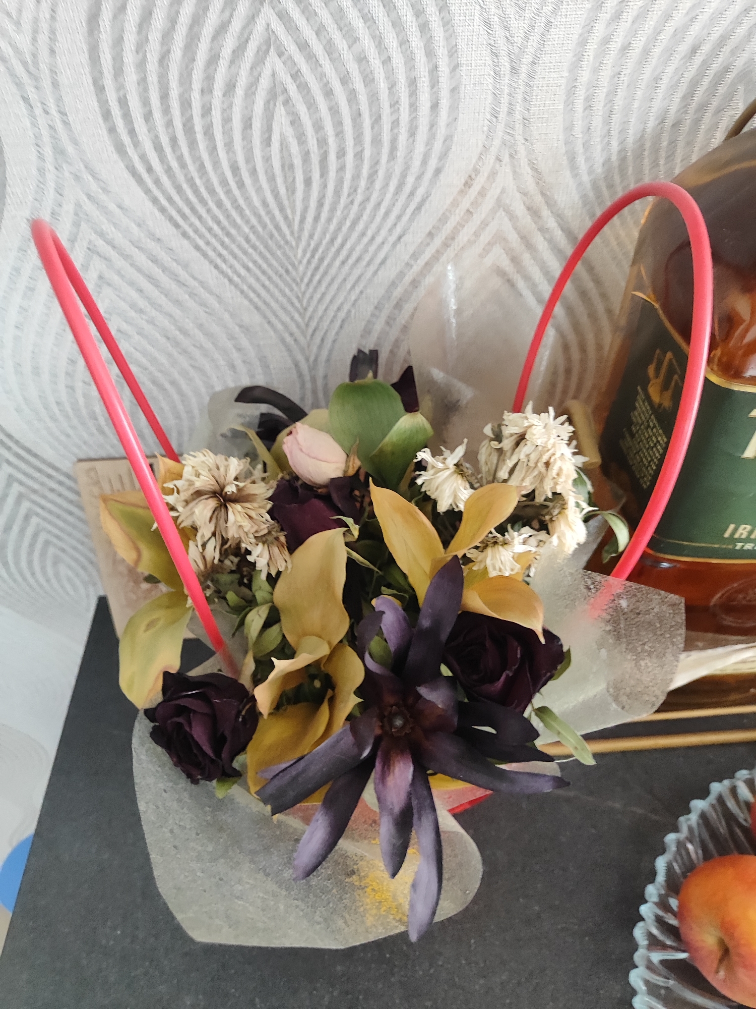 A close-up, medium shot captures a bouquet of dried flowers arranged in a basket with red handles. The bouquet features various dried blossoms, some a pale tan and others a deep purple, interspersed with dried golden leaves and a single pale pink rosebud. The basket itself appears to be made of a textured, light-colored material, possibly fabric or paper. Behind the bouquet, a bottle of amber liquid, likely whiskey, stands prominently. The background consists of a white, textured wallpaper with a repeating leaf-like pattern. The scene is set on a dark, flat surface, possibly a table, with a small glass dish containing two pieces of fruit, possibly apples, visible in the lower right corner. The lighting appears to be natural, coming from the left side of the frame, casting soft shadows.