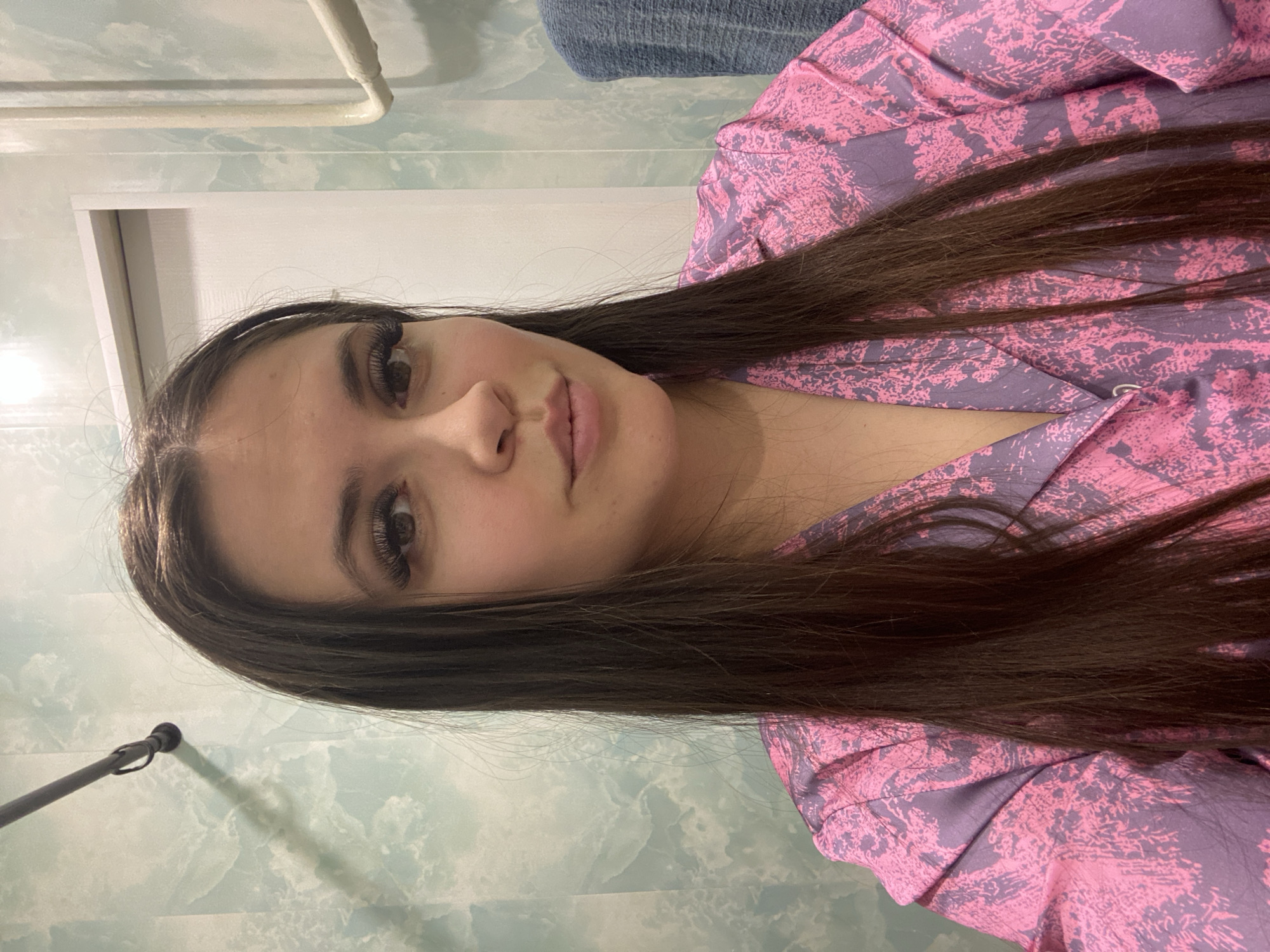 A young woman with long, dark hair is the subject of this portrait. She is wearing a pink and purple patterned garment, likely pajamas or a robe. Her skin is fair, and her makeup is apparent, particularly her eyelashes.
The background suggests an indoor setting, possibly a bathroom, given the mottled blue and white wall and the presence of a chrome rod fixture at the upper left corner. A white doorway is visible on the left.
There is no information in the image to determine its location as the city of Chaikovsky, Russia.