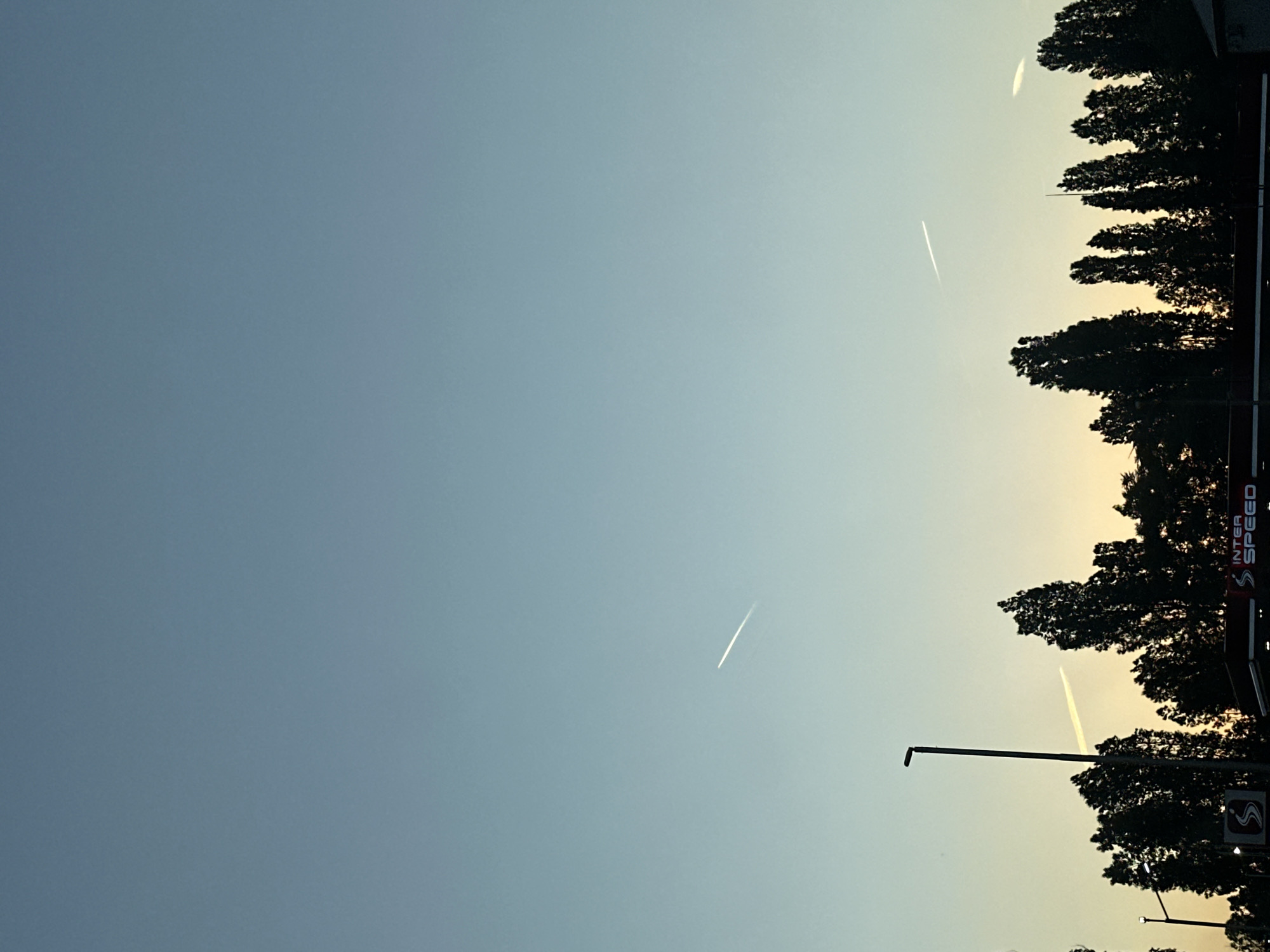 The image shows a predominantly clear sky with contrails visible, transitioning from blue to a warm, yellowish hue near the horizon. The lower part of the image contains silhouettes of trees and street lights, with a gas station sign partially visible on the right. The sign displays "INTER SPEED". This is probably taken on the outskirts of Sofia, Bulgaria.