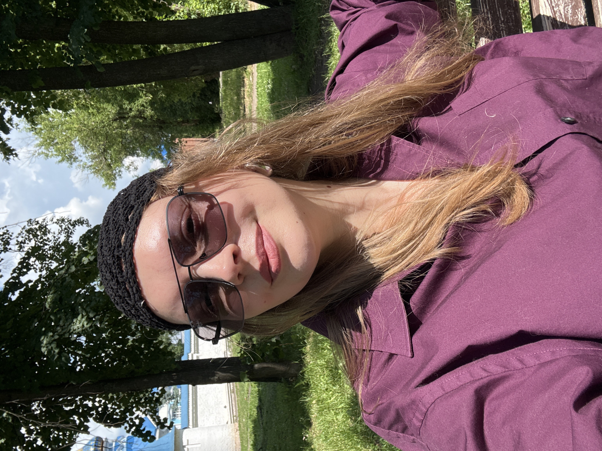 The image is a selfie of a woman outdoors, likely in a park or public space, on a sunny day. She is wearing a dark crocheted cap, large sunglasses with dark lenses, and a deep purple collared shirt. Her long, blonde hair is visible around her face and shoulders. The background shows lush green grass, trees, and a hint of white architecture with a blue dome, possibly a church or religious building, suggesting the location is in Izhevsk, Russia. The woman appears to be relaxed and smiling faintly.