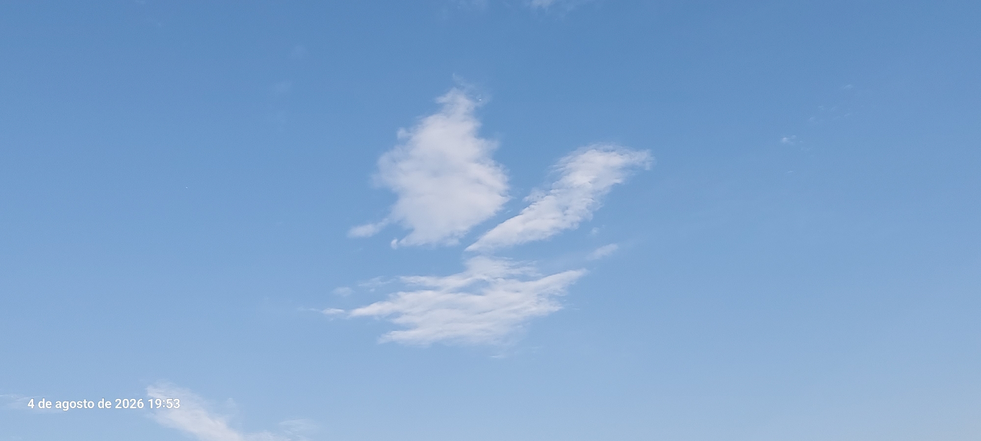 The image shows a bright blue sky with scattered white clouds. In the bottom left corner, text reads "4 de agosto de 2026 19:53", indicating the date and time the photo was taken. The scene appears to be an outdoor setting with clear weather.