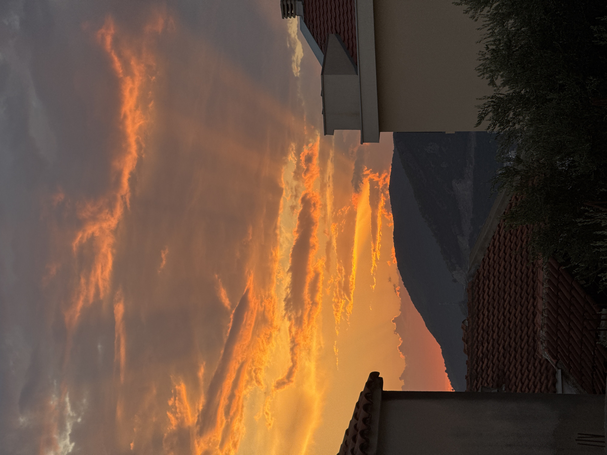 The image captures a vibrant sunset over the town of Trap, Montenegro, as seen from an elevated perspective. The sky is ablaze with fiery orange and yellow hues, painted across dramatic clouds. Rays of sunlight pierce through the clouds, casting distinct beams into the atmosphere. In the foreground, the rooftops of several houses, covered in traditional terracotta tiles, are visible. These buildings are situated amidst lush, dark green foliage. Behind the houses, a silhouetted mountain range forms a dark backdrop against the brilliantly lit sky. The scene is devoid of people or any discernible activity, presenting a serene, natural landscape at dusk.